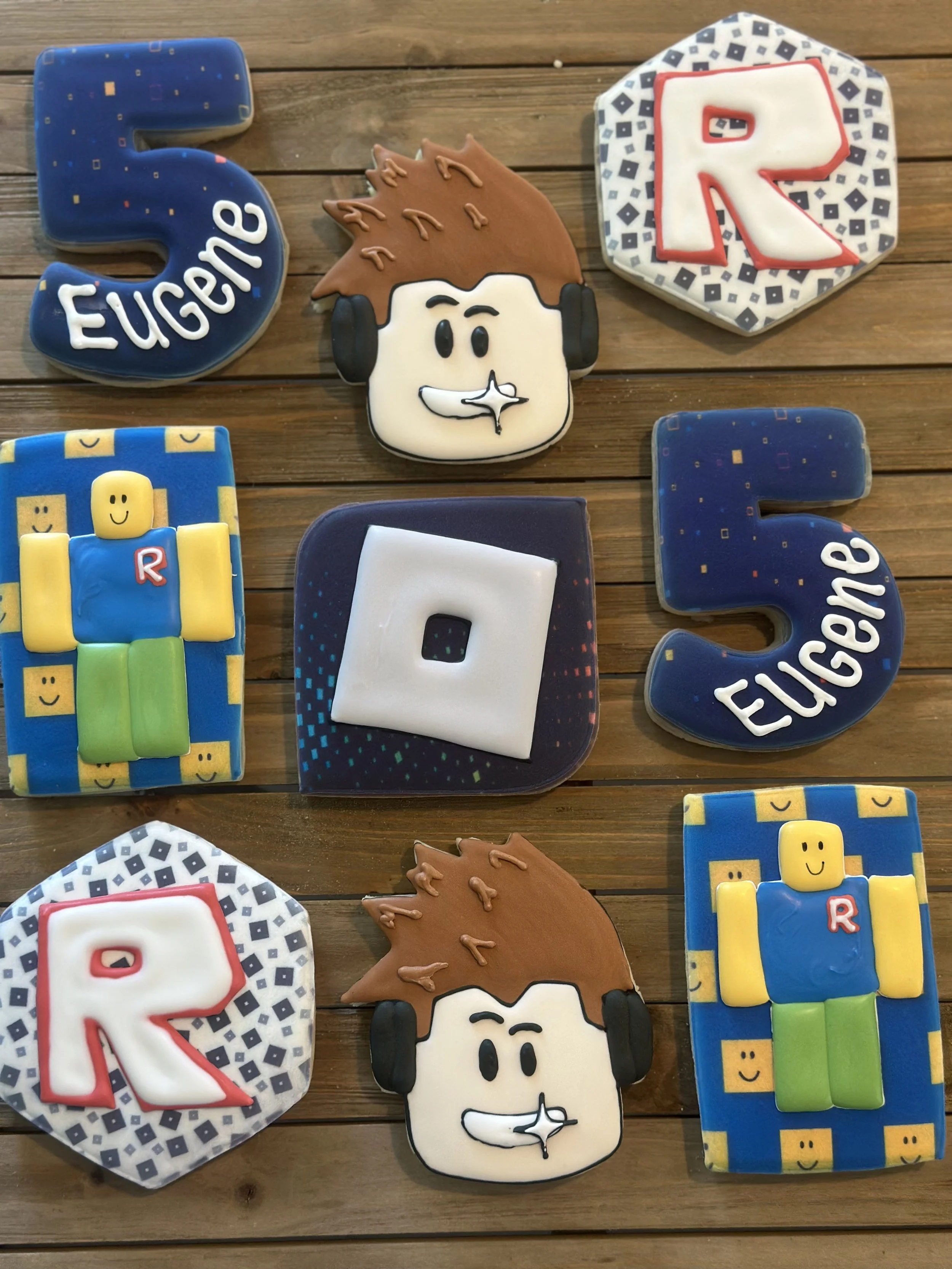 Decorative cookies with themes related to robotics and Roblox, arranged on a wooden surface. There are cookies shaped like the number 5, the letter R, a Roblox character with a blue shirt, yellow arms, and green legs, and two cookies with a face and