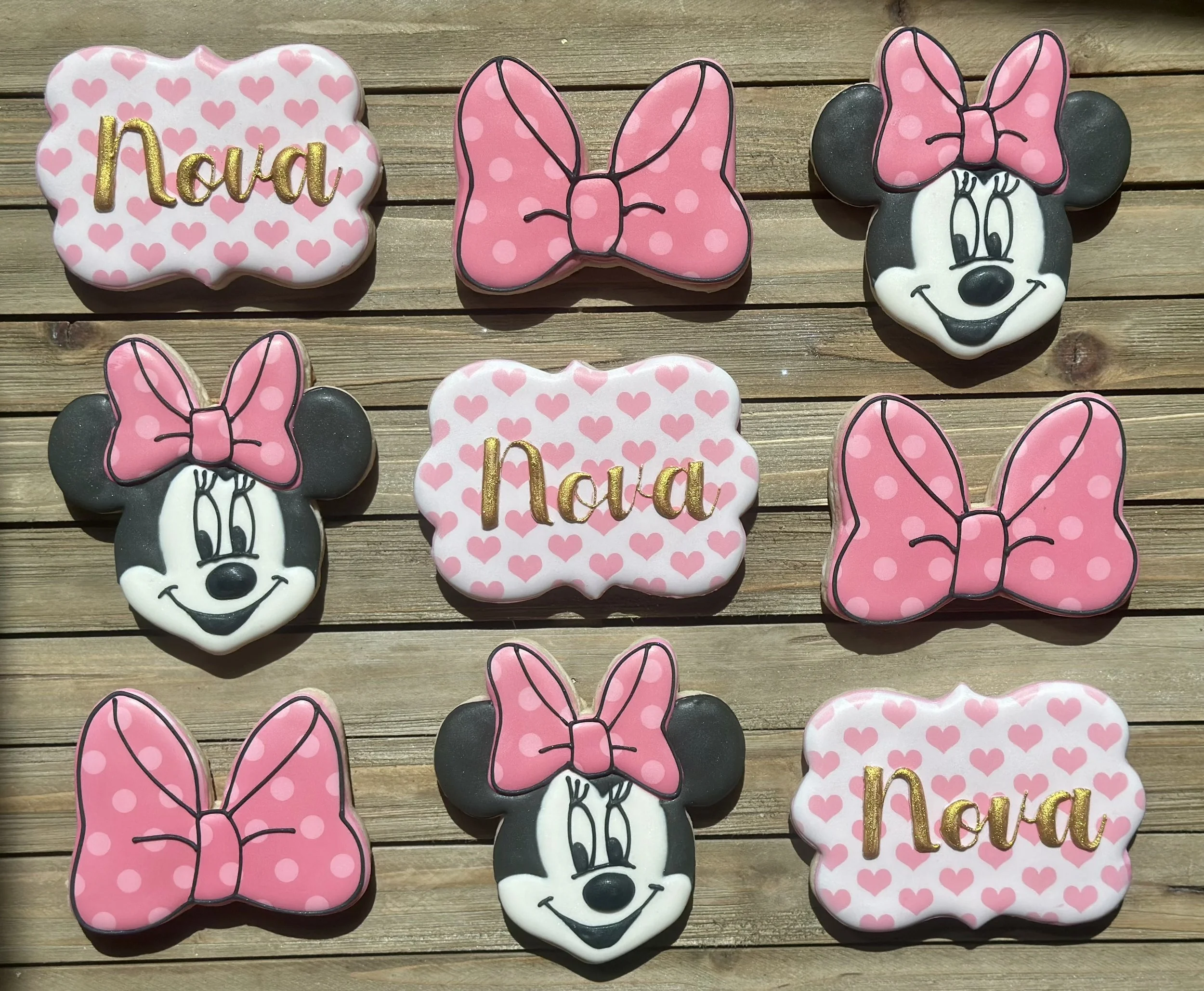 Decorated cookies arranged on a wooden surface, featuring Minnie Mouse faces with pink polka dot bows, pink polka dot bows, and white cookies with pink hearts and the name Noah in gold lettering.