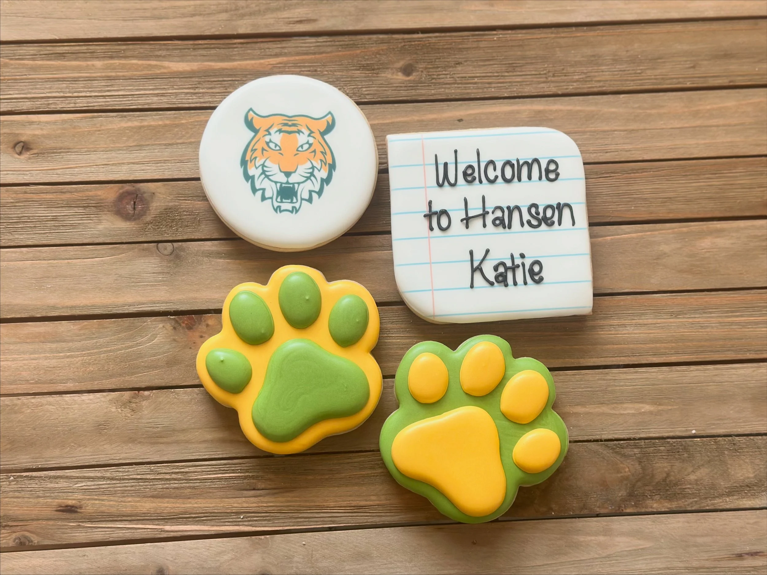 Decorative cookies on a wooden surface, one with a tiger face, two with paw prints, and a notebook-shaped cookie with the message 'Welcome to Hansen Katie' written on it.