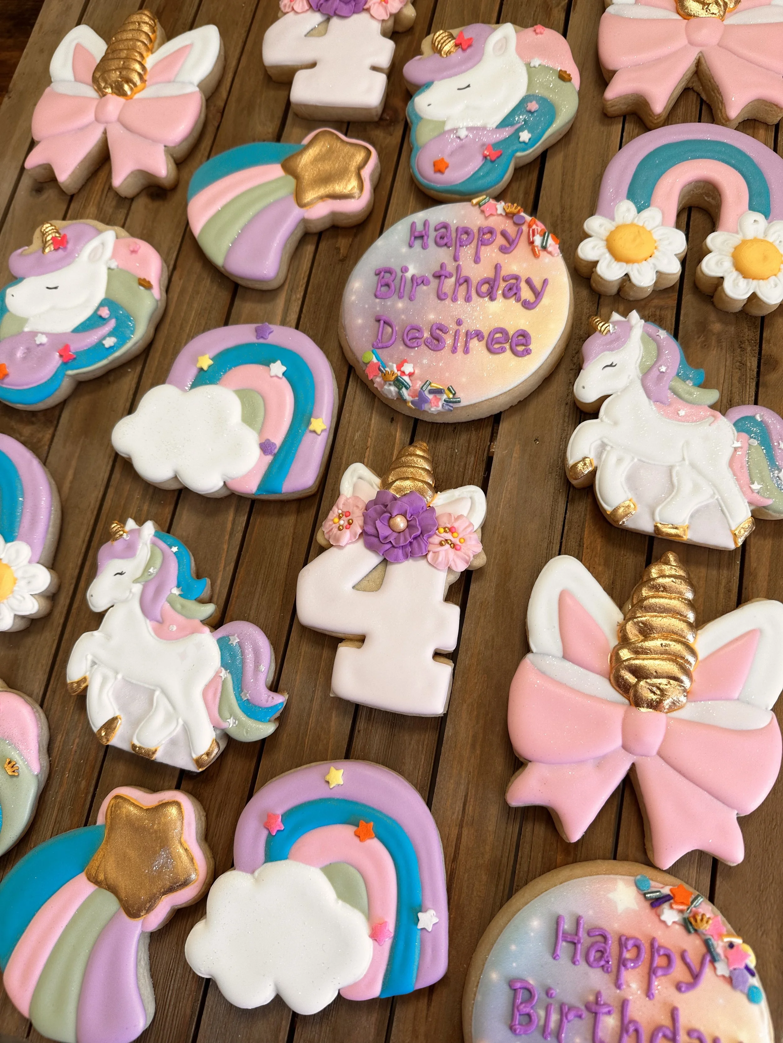 Colorful unicorn and rainbow themed birthday cookies on a wooden table, including a cookie with the message 'Happy Birthday Desiree' and various decorative cookies shaped as unicorns, rainbows, stars, clouds, and bows.