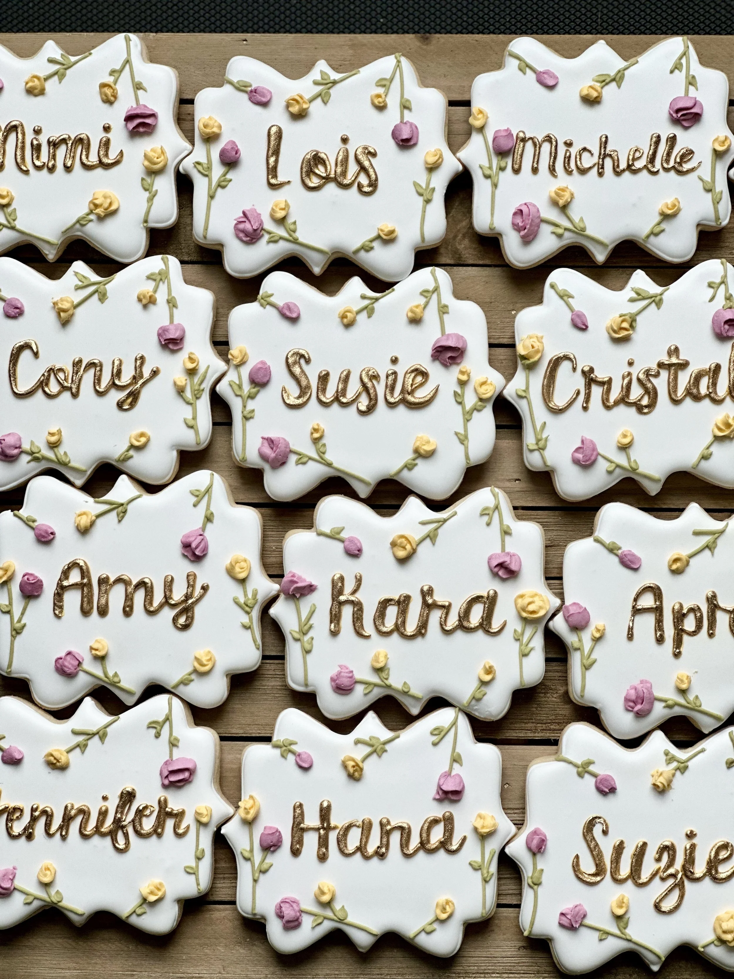Decorated cookies with white icing, each inscribed with a different name in gold or brown icing, decorated with small pink and yellow flowers and green leaves.