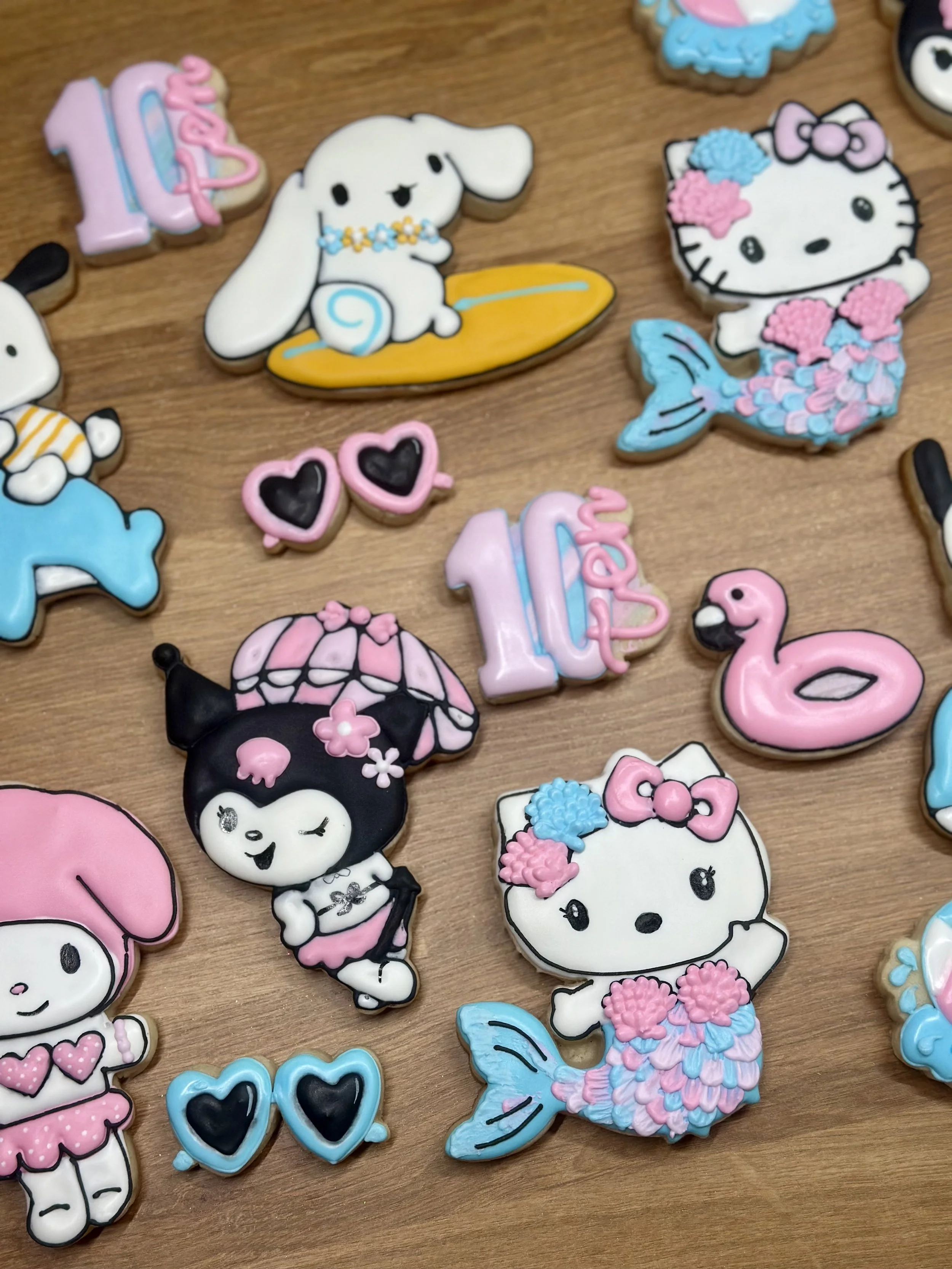 Decorated cookies shaped like Hello Kitty, a dog, a girl, a flamingo, hearts, and the number 10 with pink, blue, and black icing on a wooden surface.