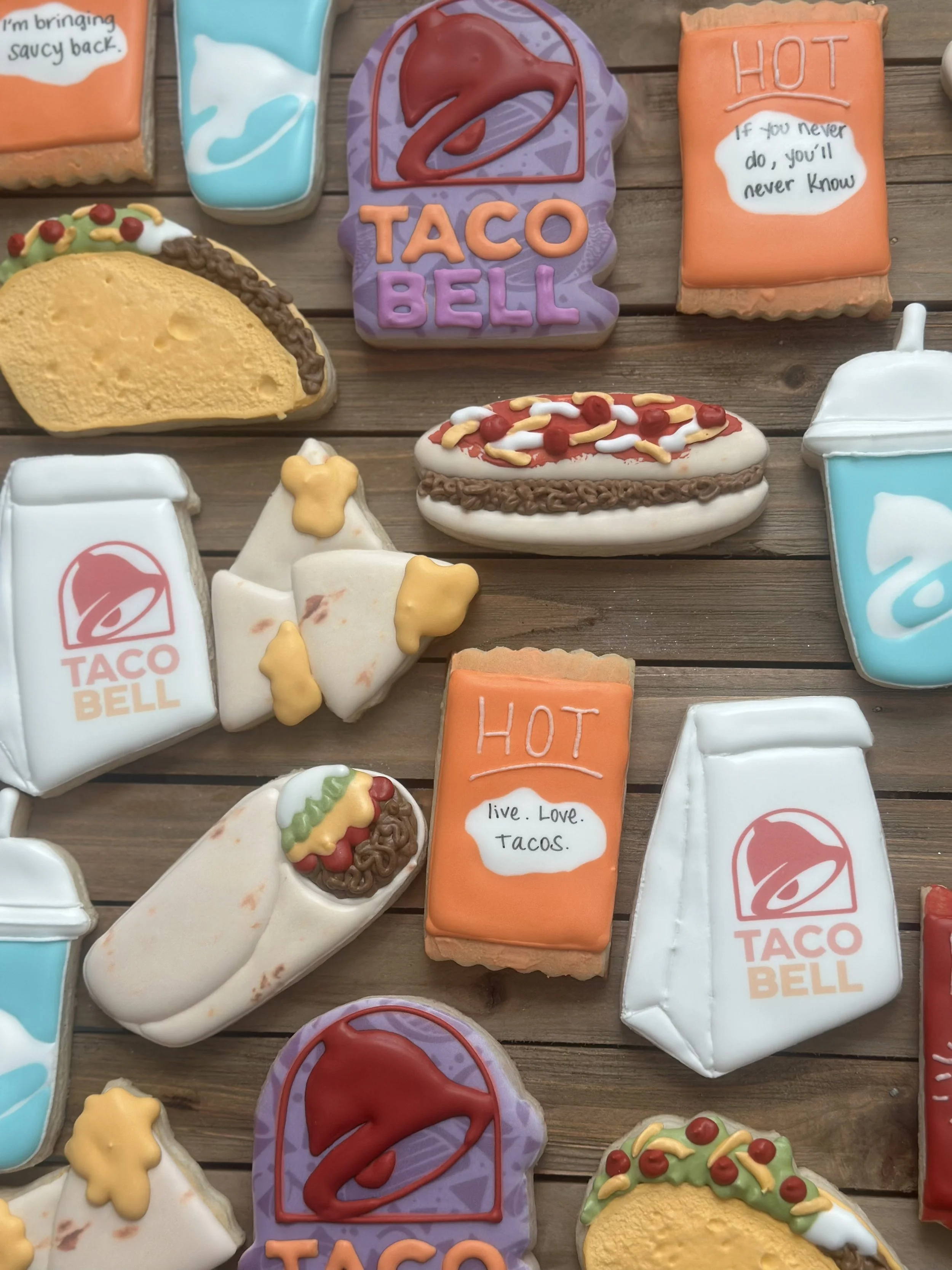 Assorted cookies with Taco Bell branding and slogans, decorated with taco designs, soda cups, and hot sauce packets on a wooden surface.