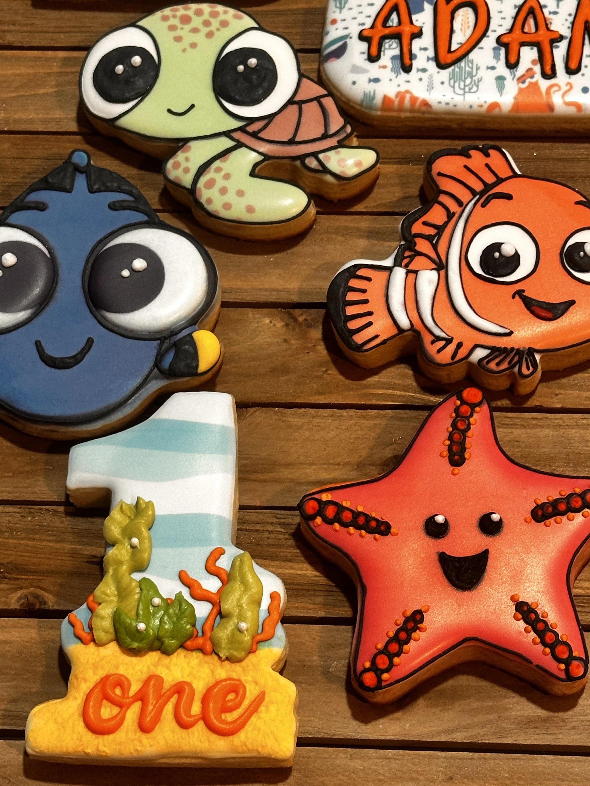 Decorative cookies with ocean and starfish themes, including a turtle, fish, starfish, and a 'one' cookie with seaweed illustrations, on a wooden surface.