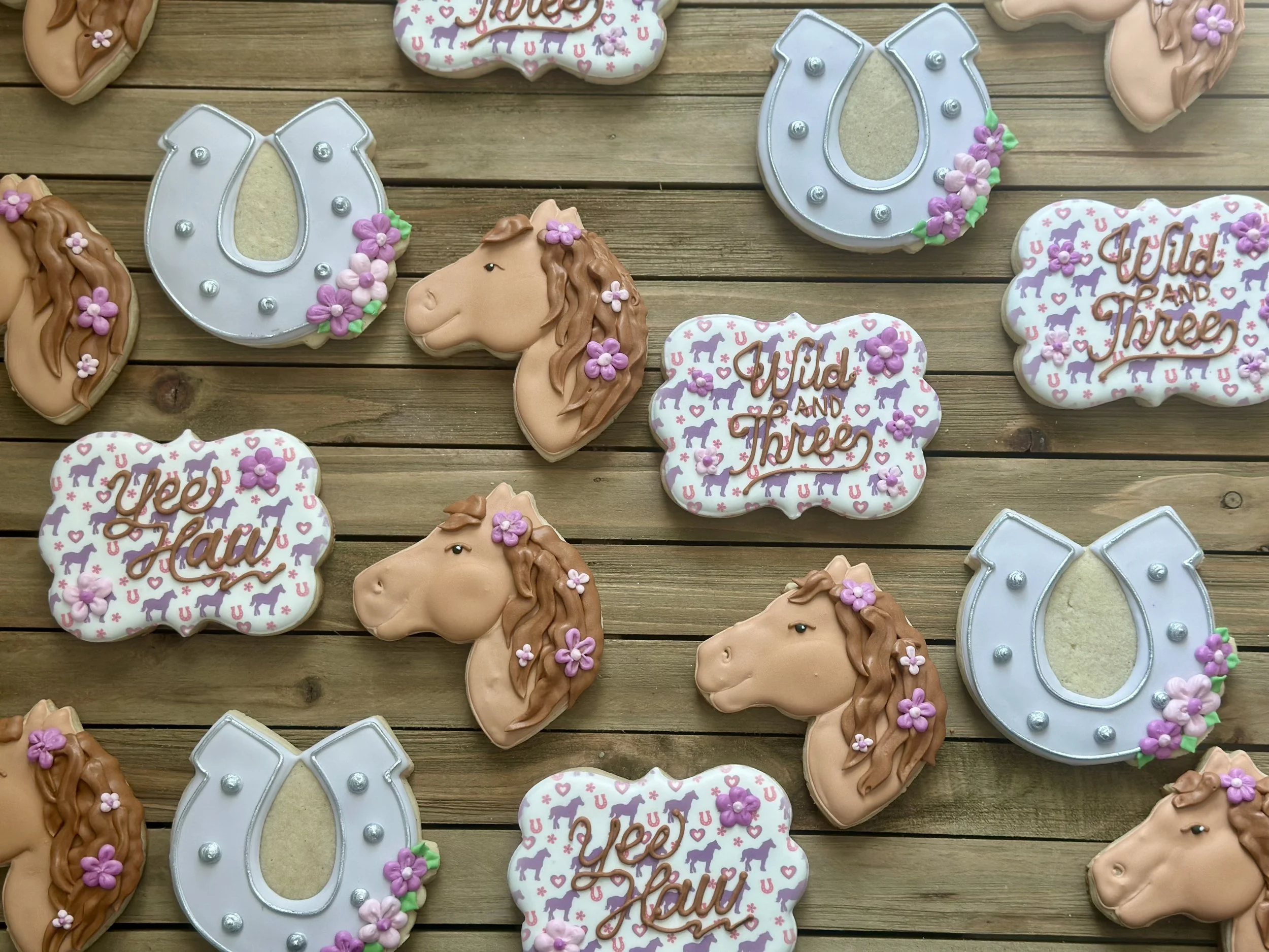 Decorative cookies with cowboy hats, horse heads, and speech bubbles with the text 'Yee Haw' and 'Wild and Three', decorated with pink and purple flowers, arranged on a wooden surface.