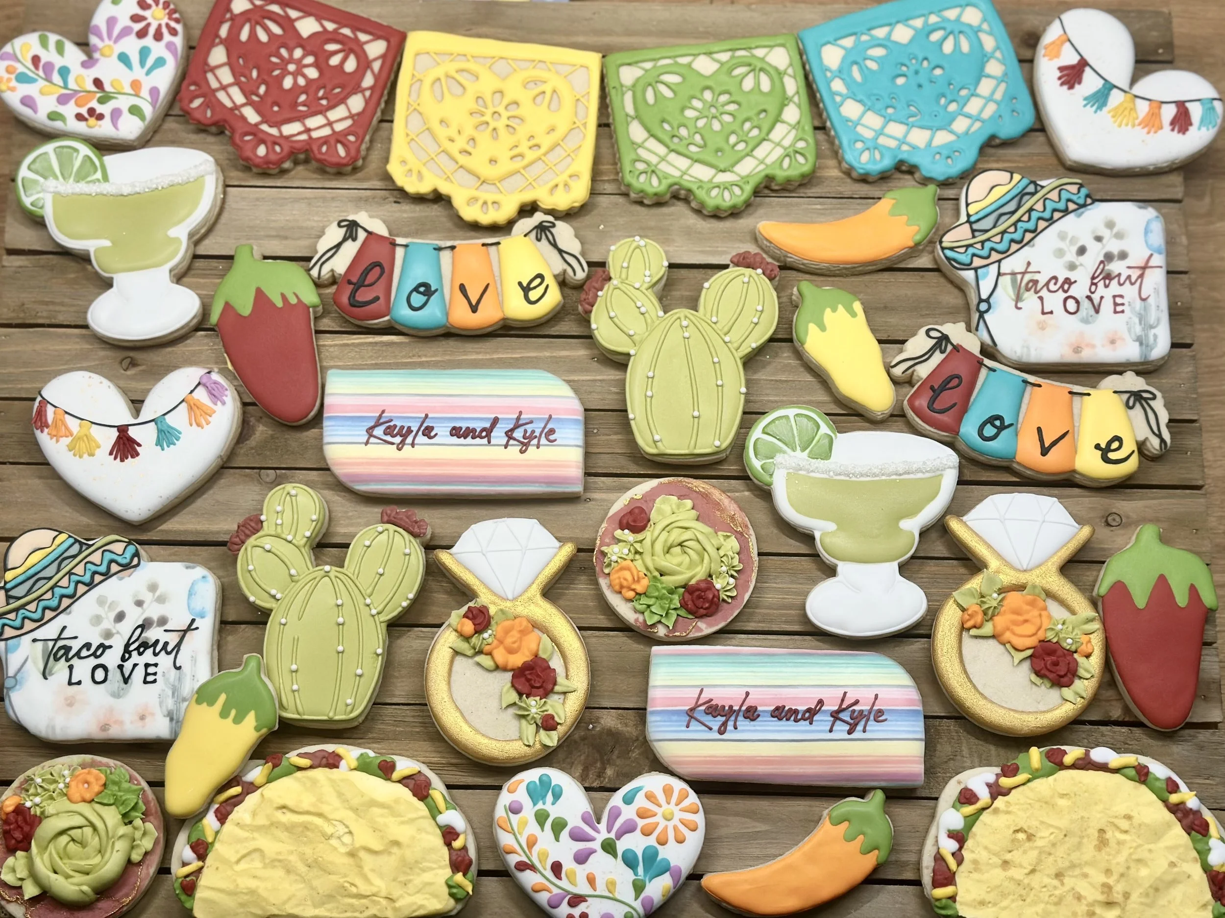 Decorated sugar cookies with various shapes and designs, including hearts, cacti, chili peppers, sombreros, margarita glasses, and the words "love" and "taco bout love" on colorful backgrounds, arranged on a wooden surface.