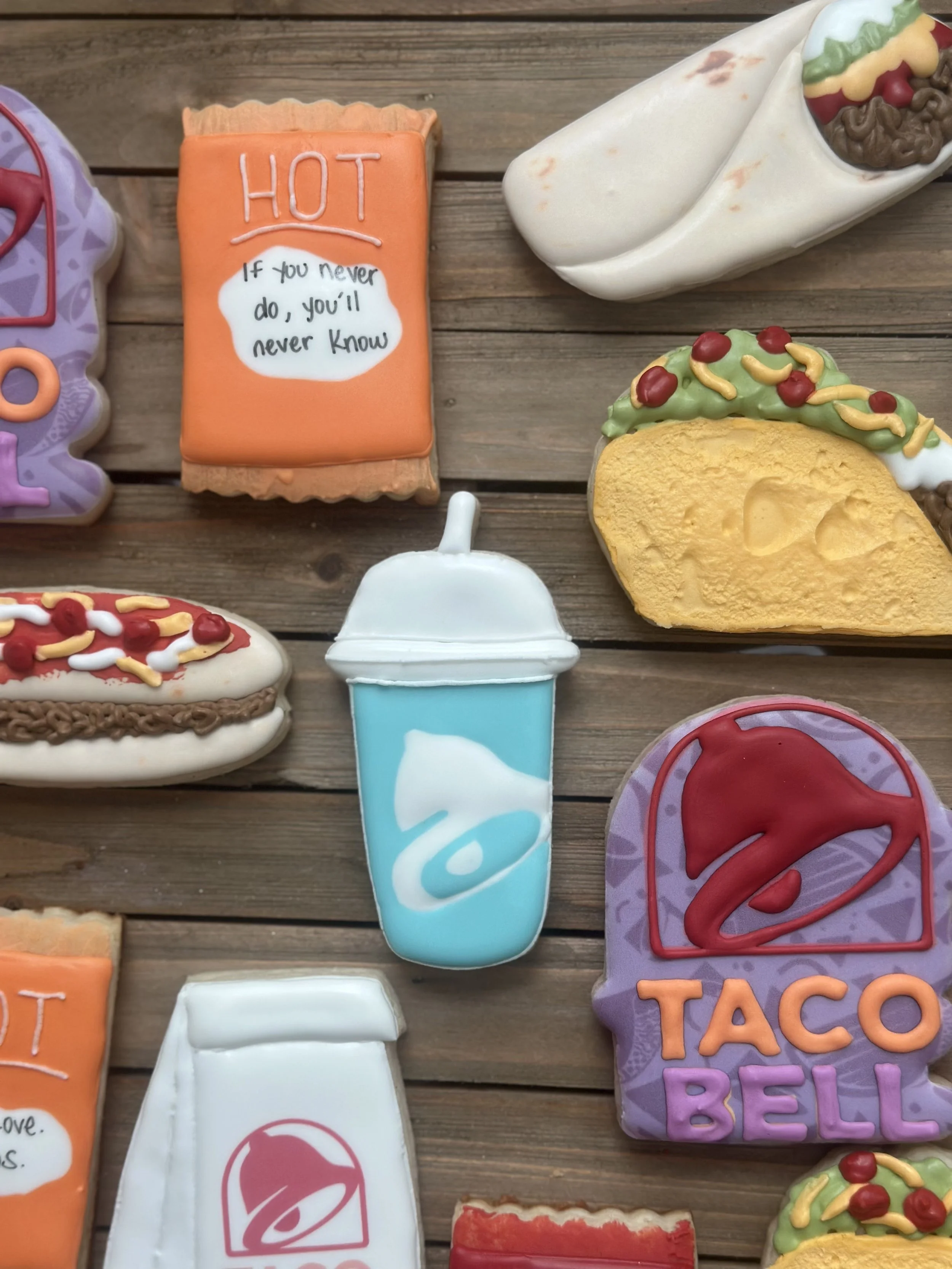 Decorative cookies shaped like a soda cup, taco bell logo, hot sauce packet, and various taco-shaped treats on a wooden surface.