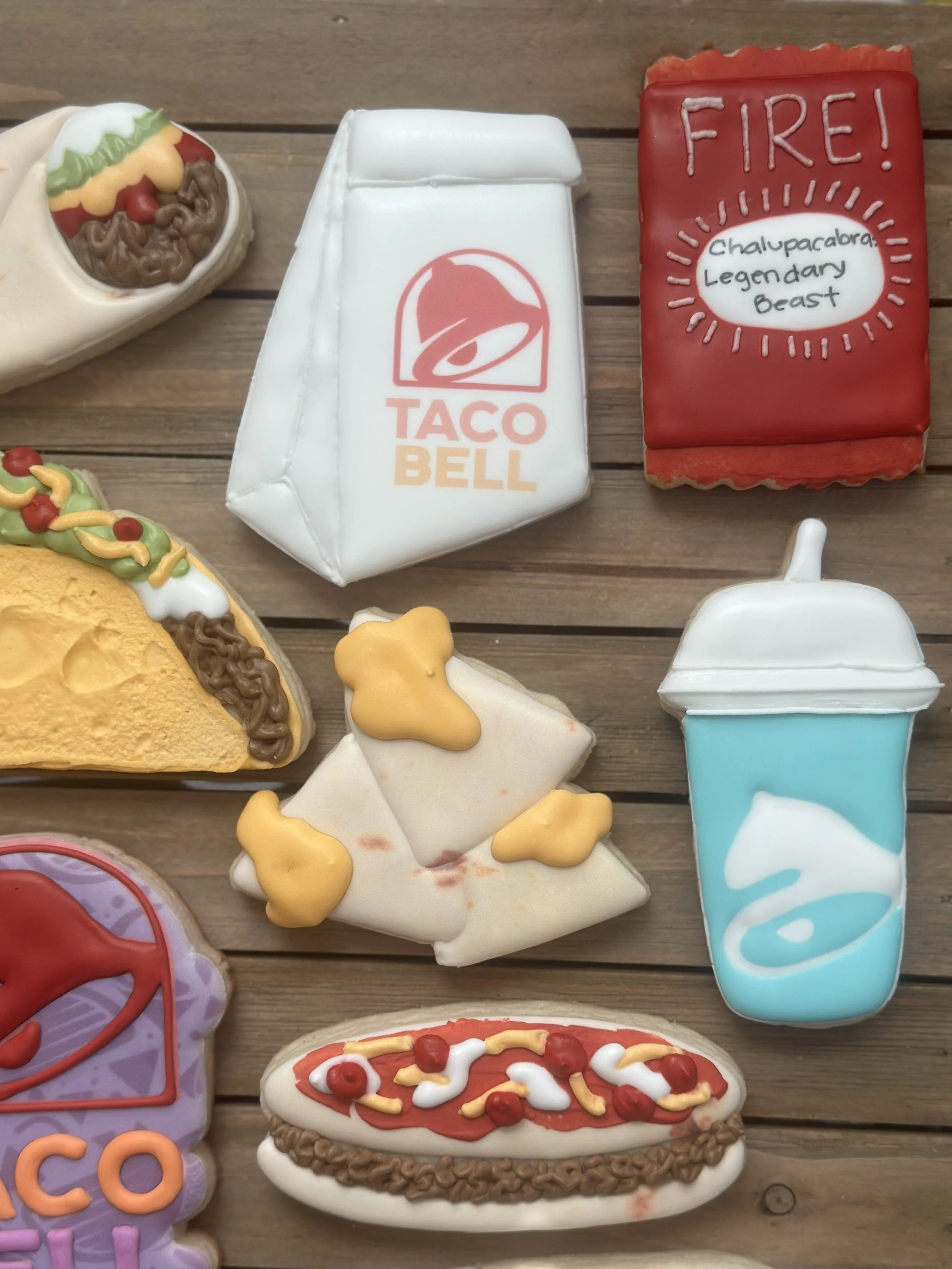 Decorative cookies shaped like Taco Bell items, including a box, a drink cup, a taco, a burrito, and a hot dog, all on a wooden surface.