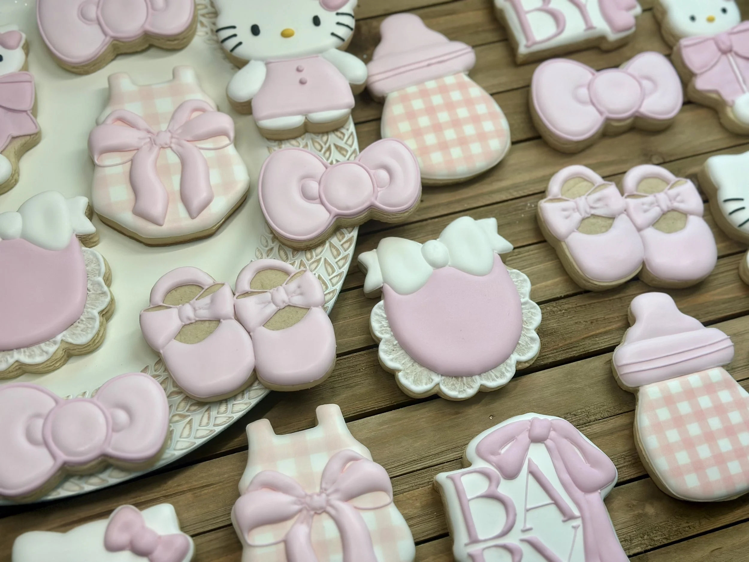 Various pink, white, and light purple decorated cookies for a baby shower, including bows, baby bottles, onesies, and a Hello Kitty character, arranged on a wooden surface.