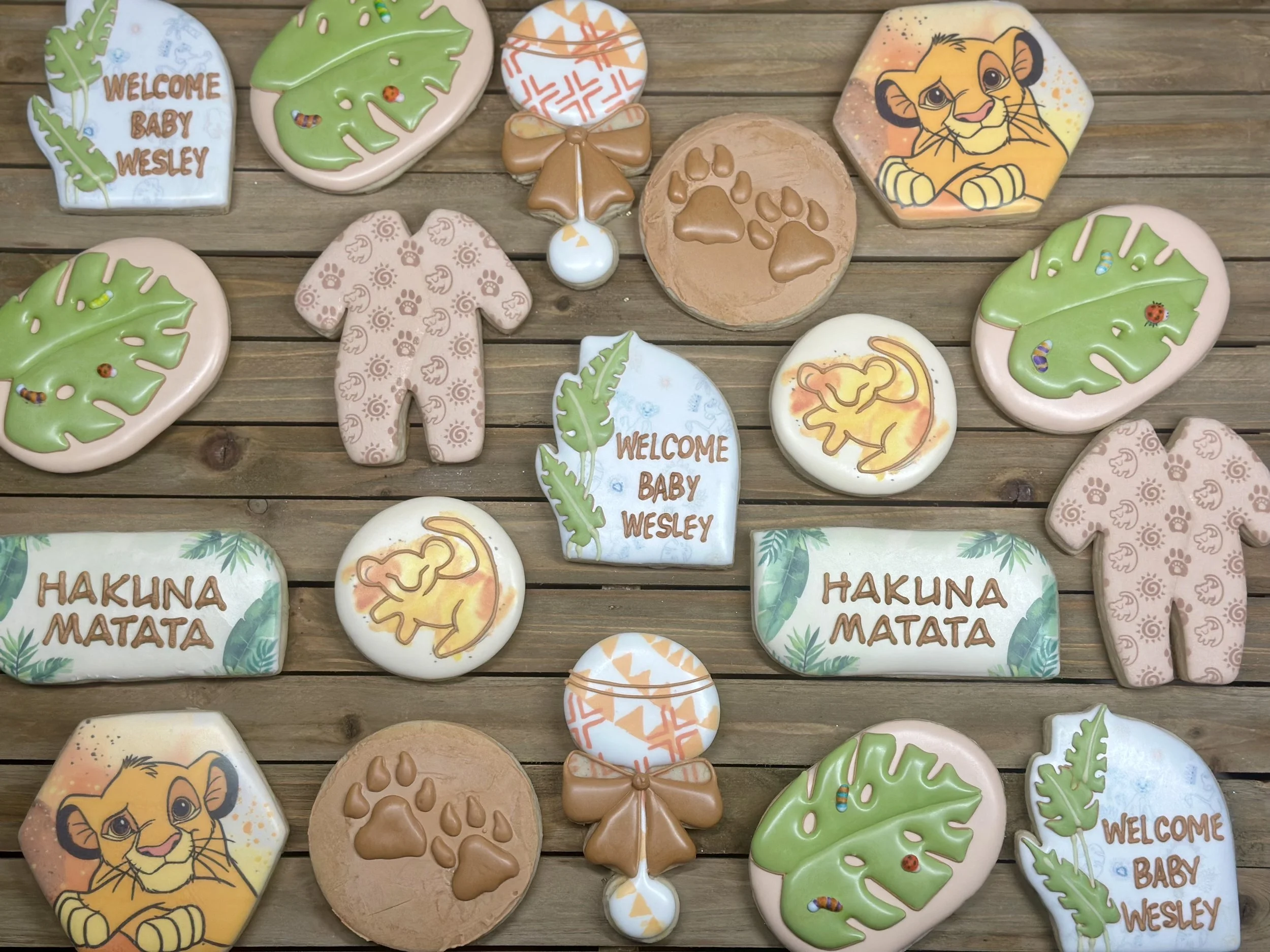 Decorated cookies with animal and baby-themed designs, including lions, paw prints, baby clothes, leaves, and messages like 'Welcome Baby Wesley' and 'Hakuna Matata,' arranged on a wooden surface.