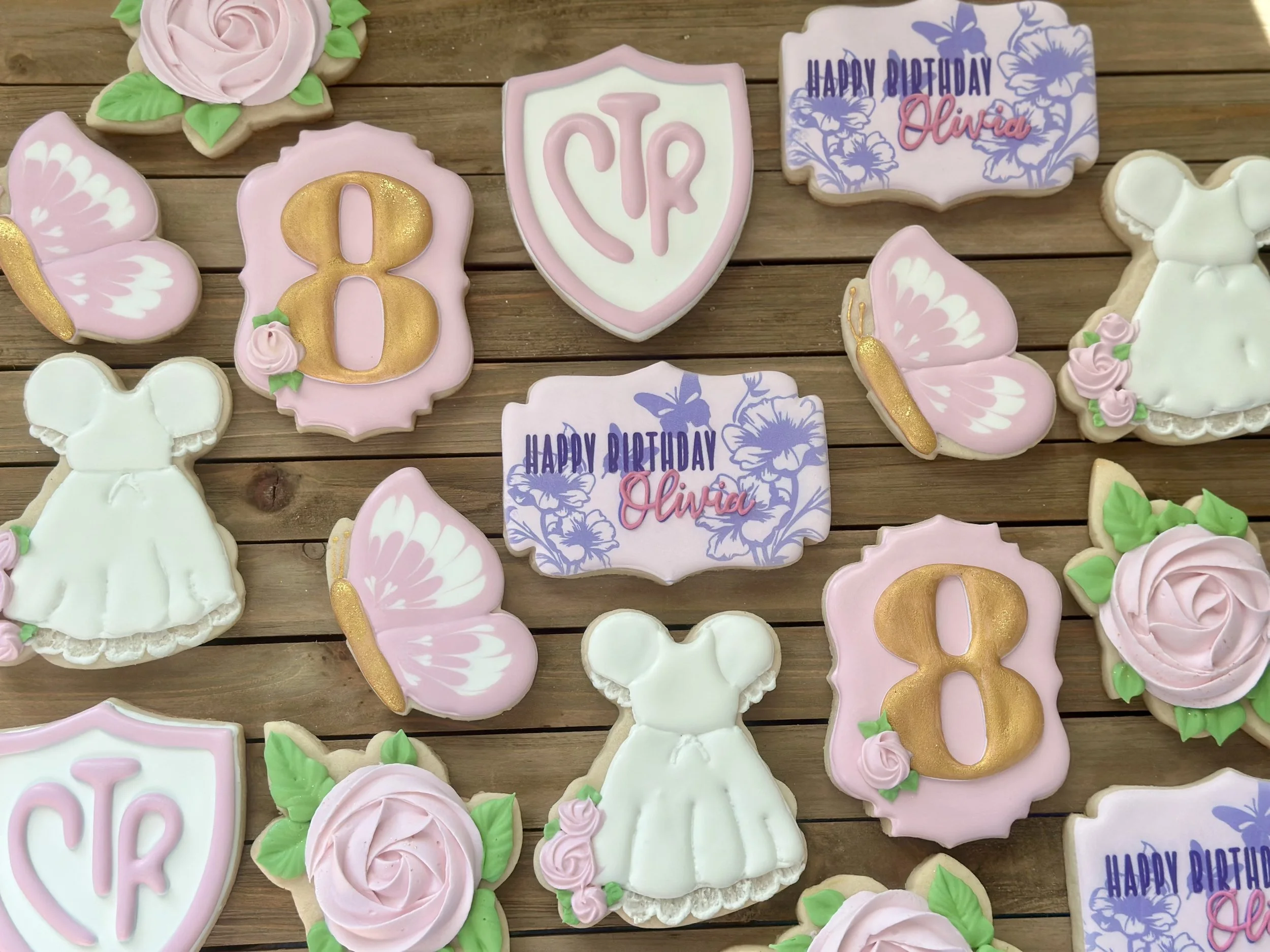 Assorted decorated sugar cookies with birthday themes, including flowers, butterflies, a princess dress, a shield with initials, and an age number, arranged on a wooden surface.