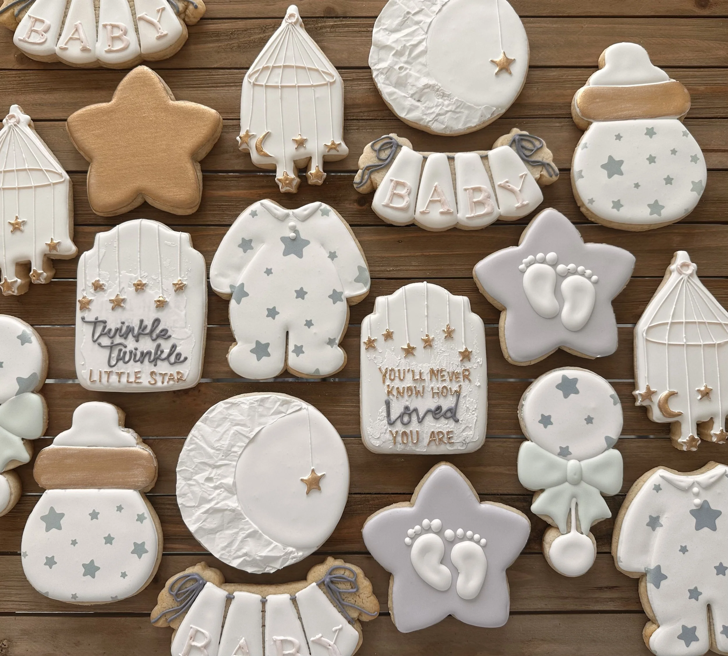 Assorted baby shower cookies with star, moon, and baby footprints designs, decorated in white, gray, and tan colors on a wooden surface.