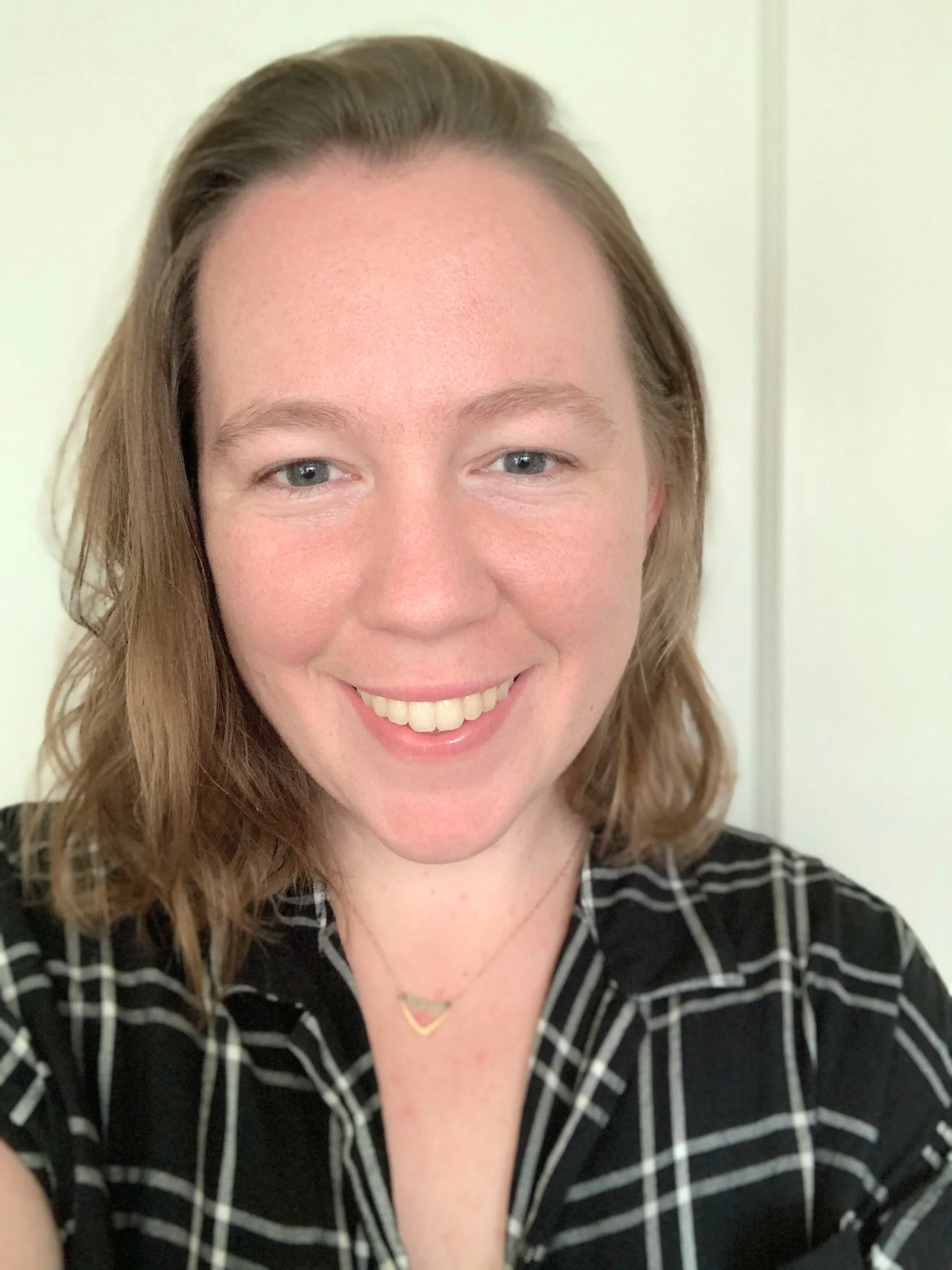 Close-up of a woman with shoulder-length light brown hair, blue eyes, wearing a black and white checked shirt, smiling, with a delicate gold necklace featuring a small triangle pendant.