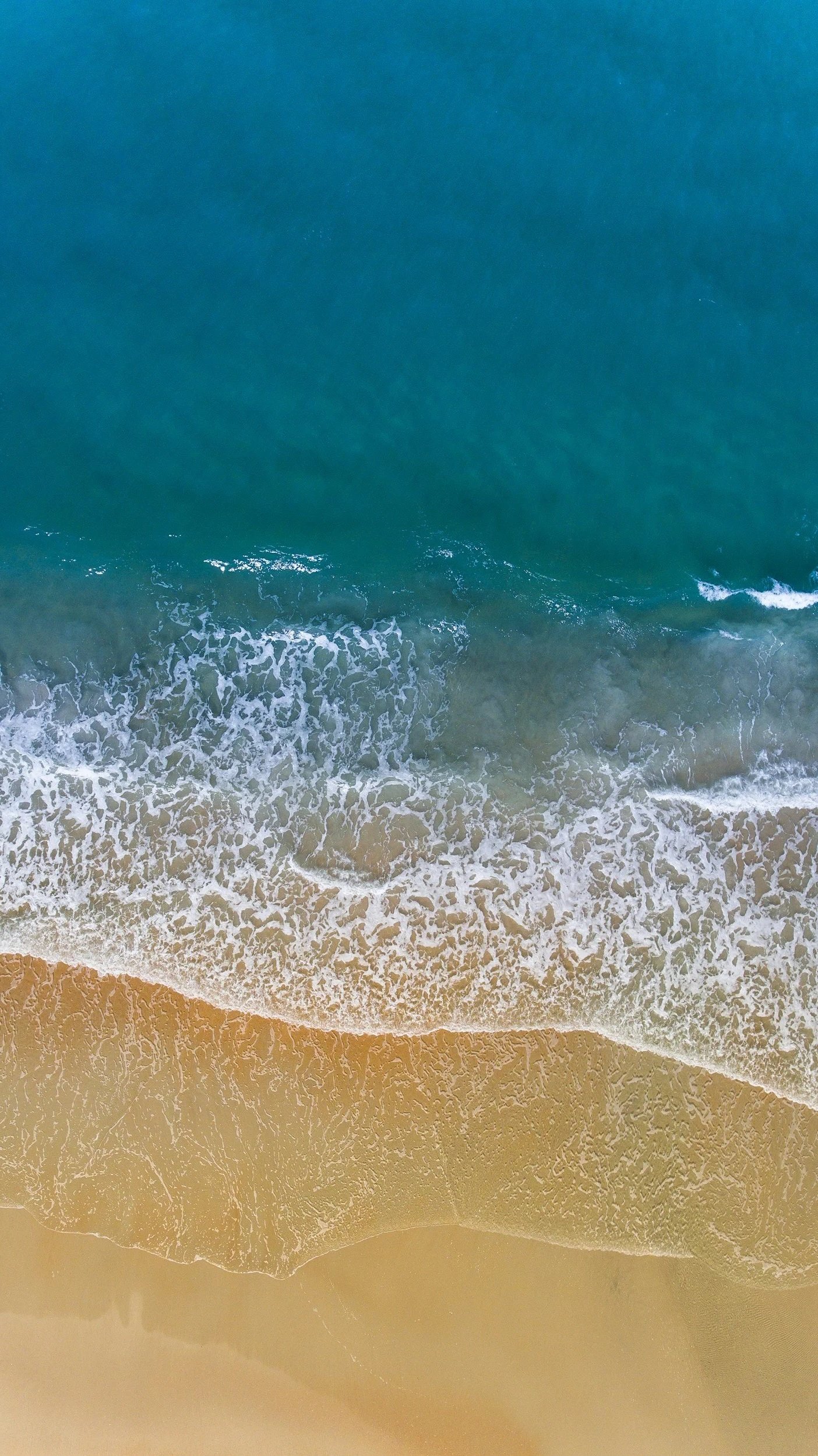 An aerial view of a beach with blue water, waves hitting the shoreline, and sandy beach area.