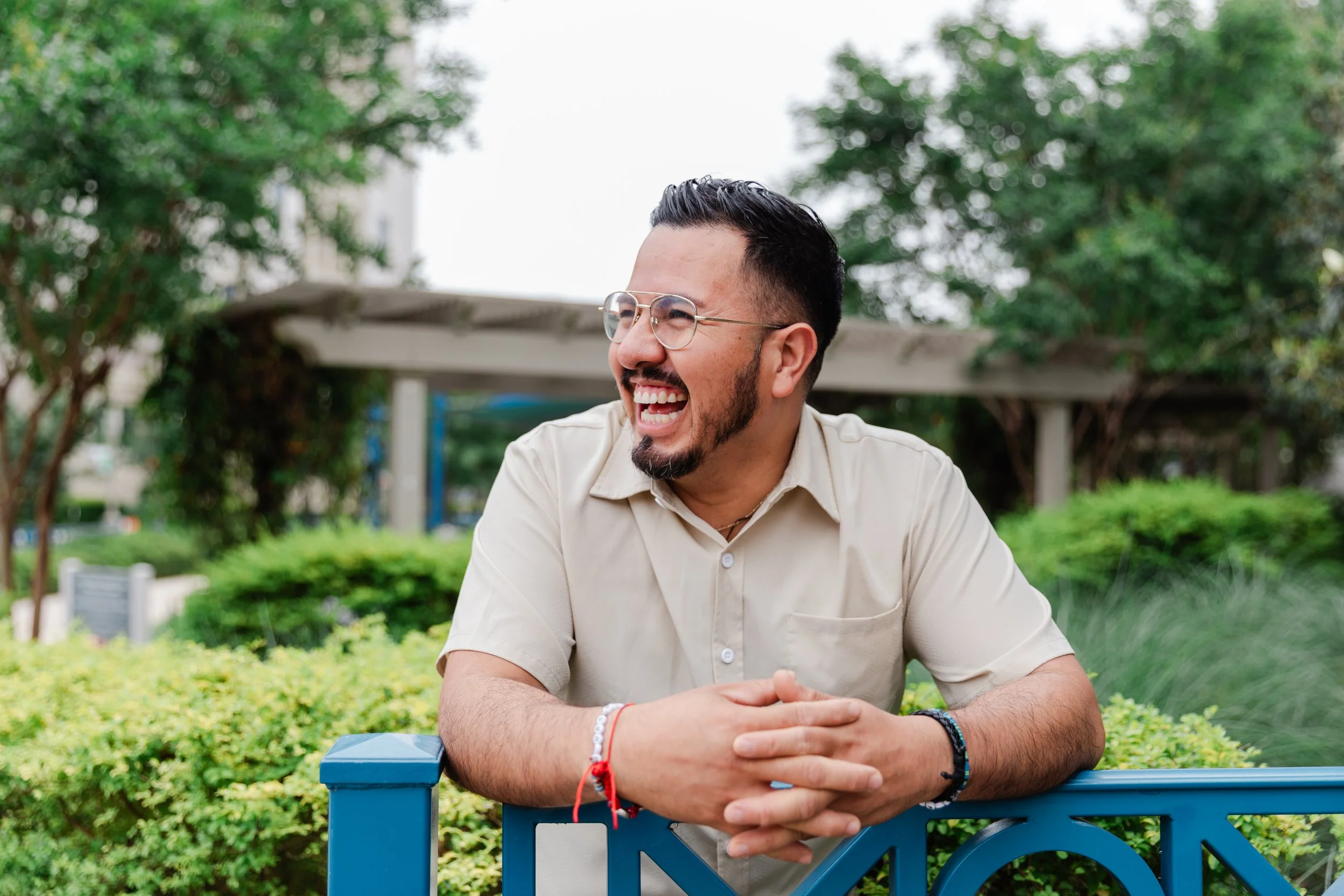 A man with glasses and a beard, smiling and laughing outdoors in a park, leaning on a blue railing with greenery and trees in the background.