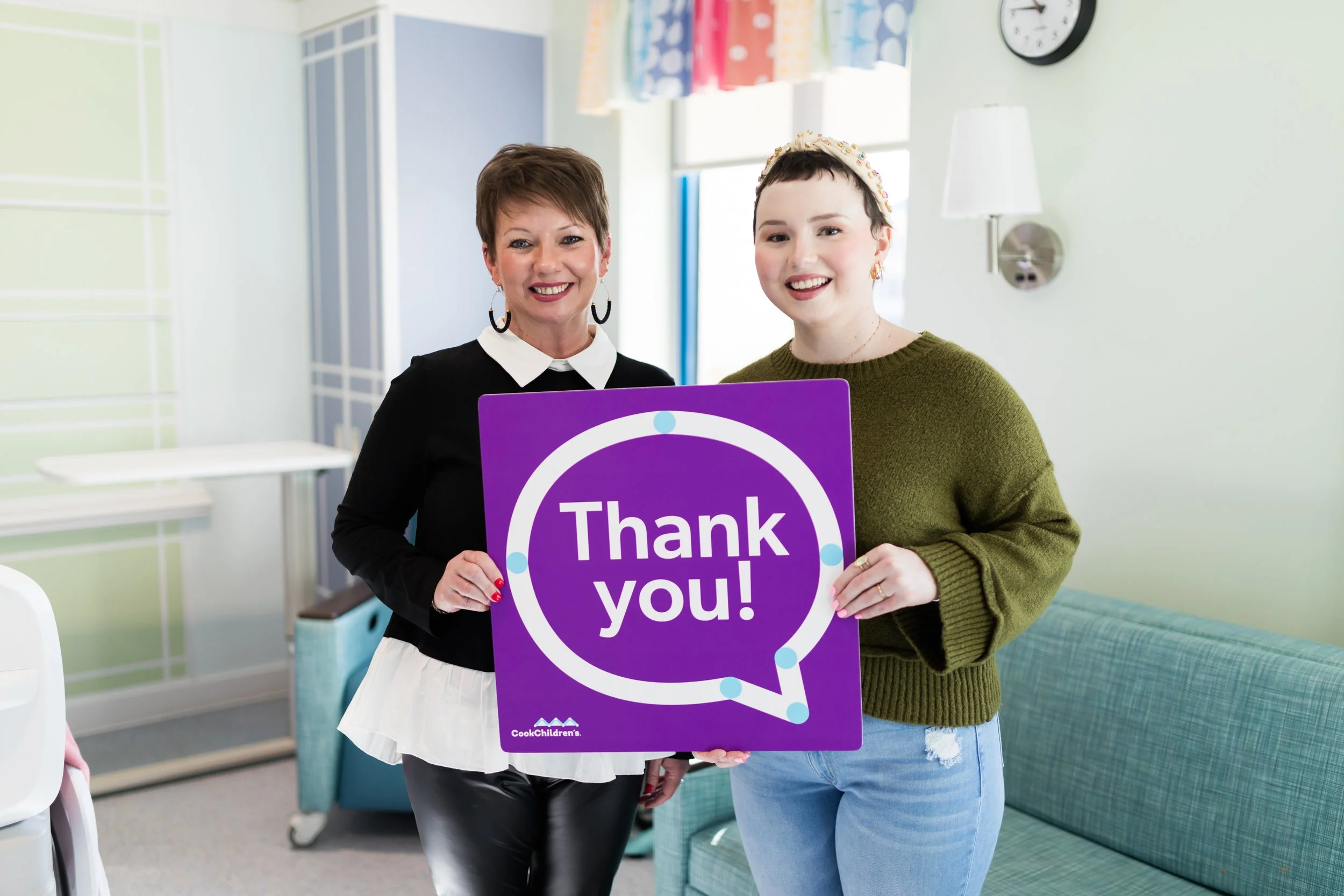 Two women holding a purple sign that says "Thank you!" in a colorful room with a green couch and a clock on the wall.