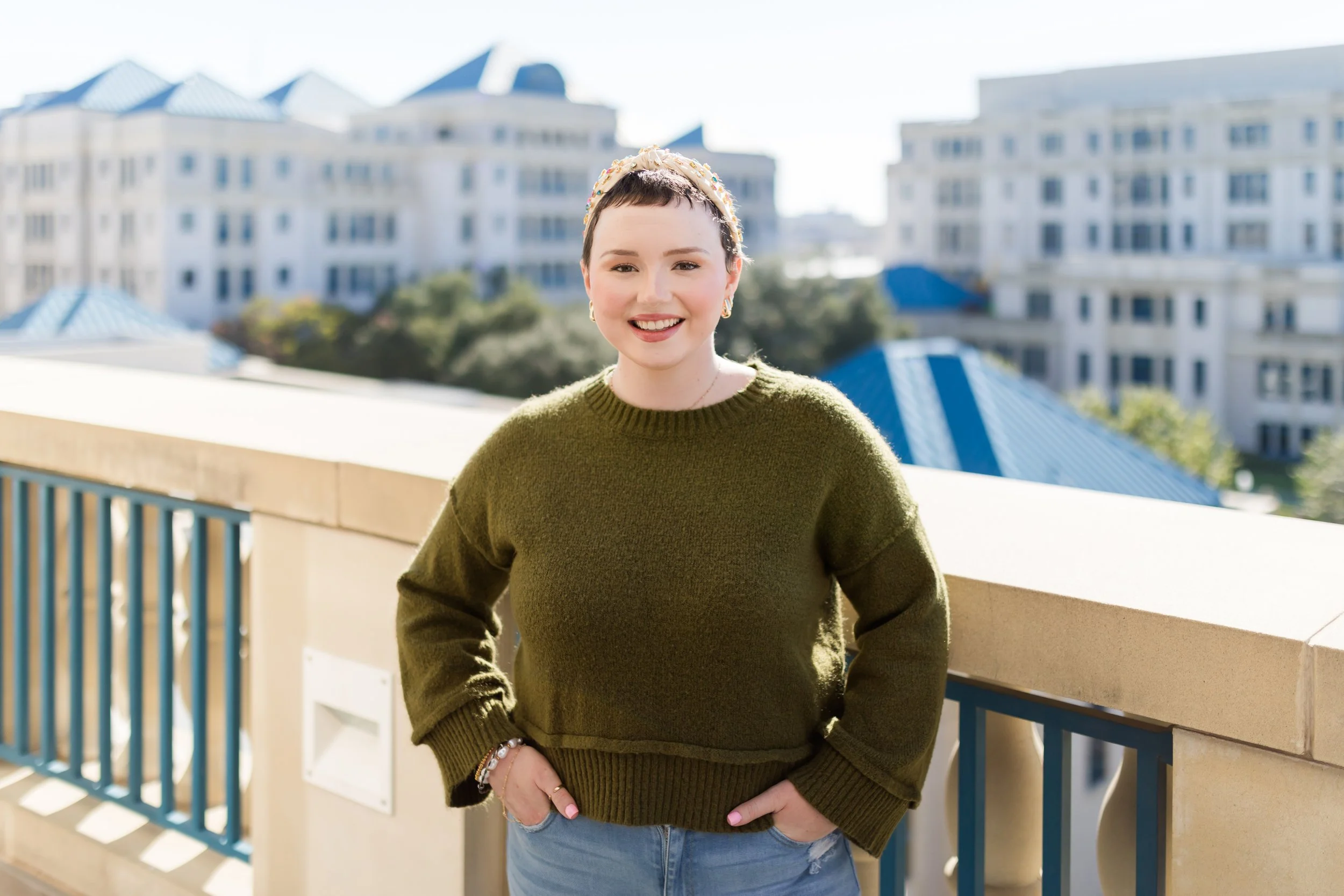 A smiling woman with short dark hair wearing an olive green sweater and jeans, standing outdoors on a balcony with a cityscape of white buildings with blue roofs in the background.