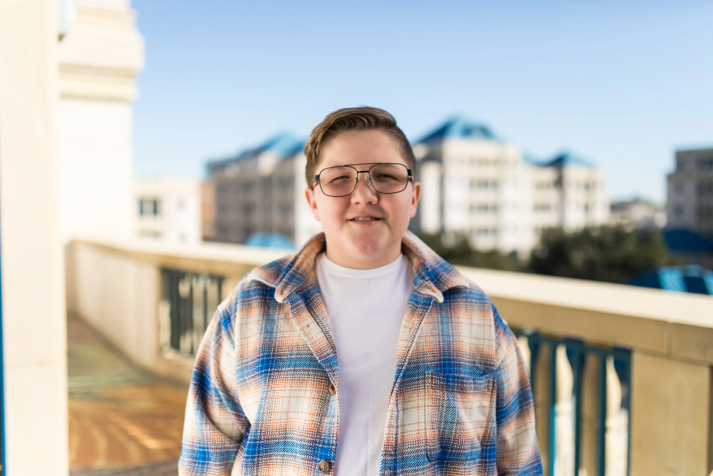 A young person standing outside on a balcony with a cityscape in the background, wearing glasses, a plaid shirt over a white t-shirt, and smiling.