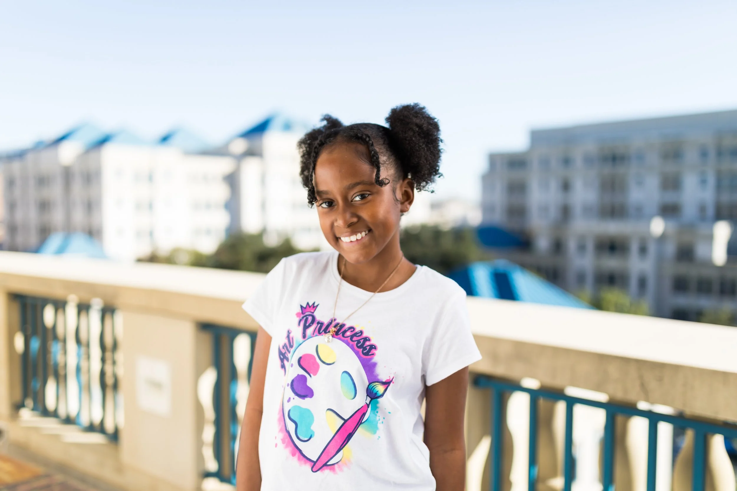 A young girl with curly hair styled into two puffs, smiling and standing outdoors on a balcony with buildings in the background, wearing a white t-shirt with a colorful art palette design and the words 'Art Princess'.