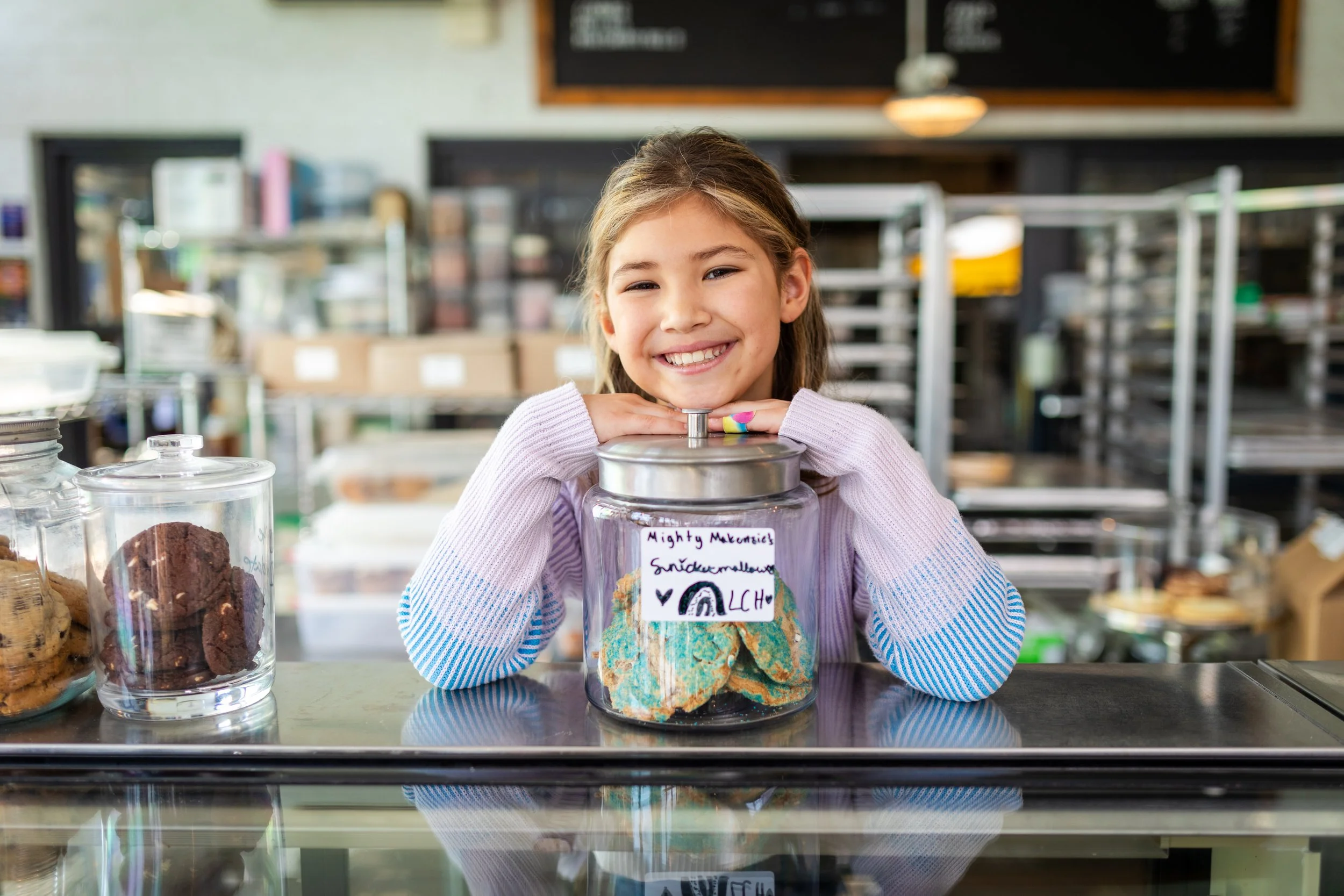 A young girl with long light brown hair smiling and resting her chin on her hands, which are placed on a glass jar filled with colorful cookies, in a bakery or cookie shop setting.