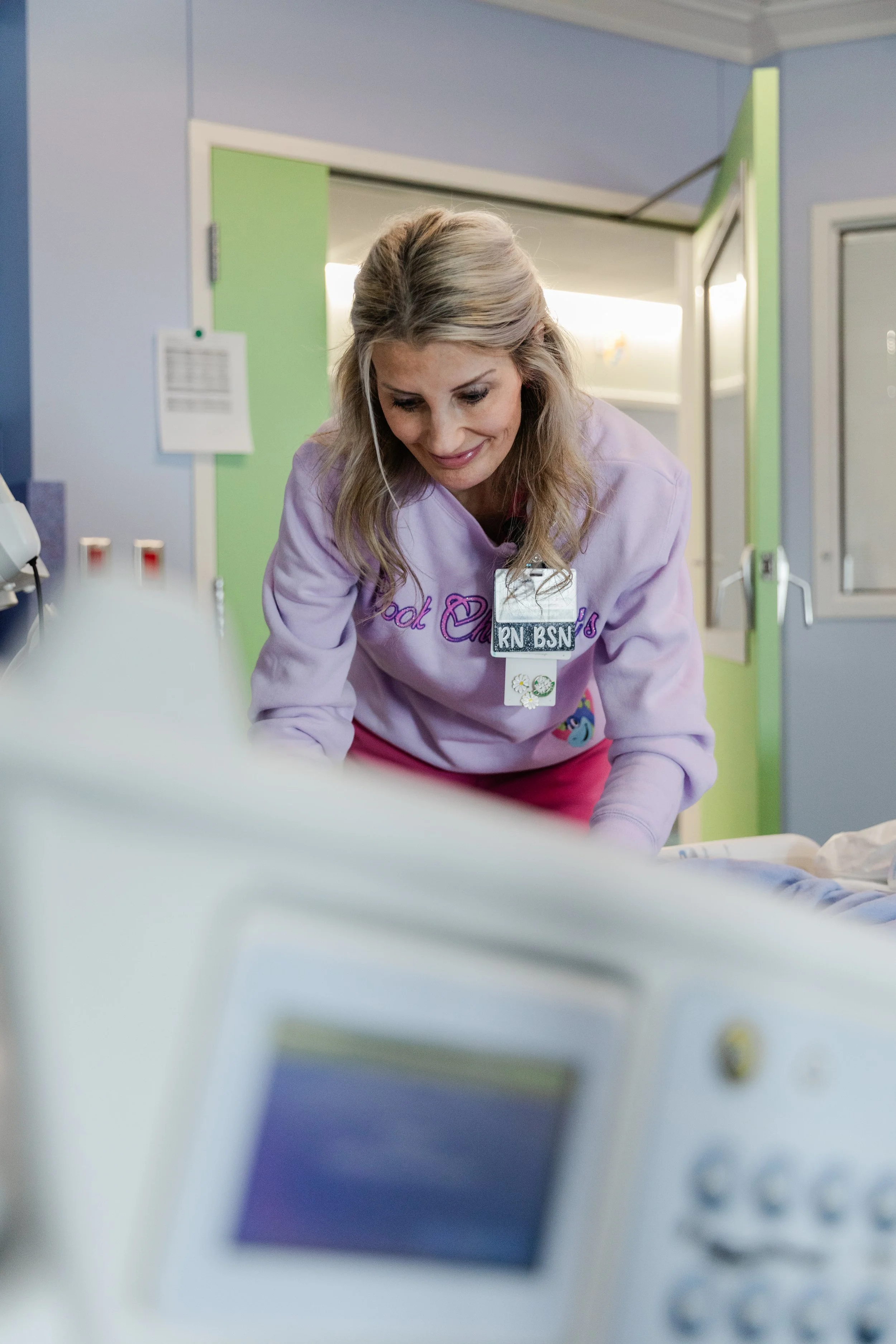 A nurse in pink scrubs with a badge reading "RN BSN" leaning over a hospital bed, smiling, in a hospital room with light blue walls and a green door.