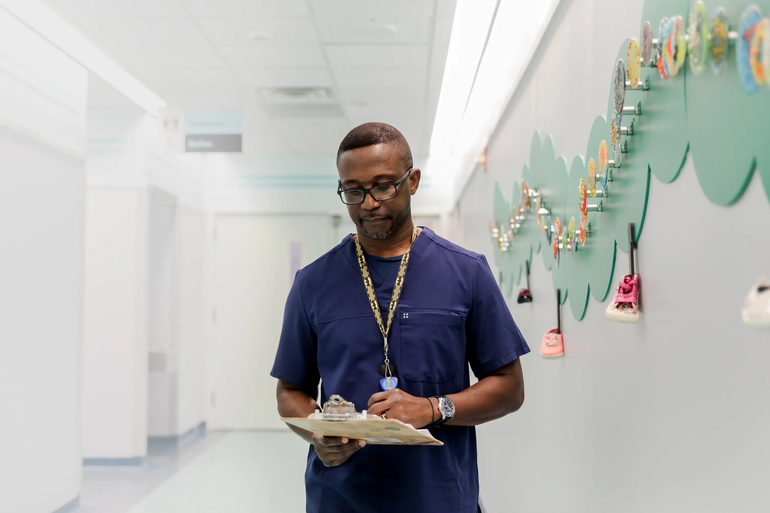 A healthcare worker wearing scrubs and glasses walking in a hospital corridor, holding a clipboard. Children’s shoes are displayed on a decoration on the wall.
