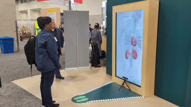 People at a science exhibition at BI Booth, looking at a large digital display of a human circulatory system, with a green carpeted area and informational booths in the background.