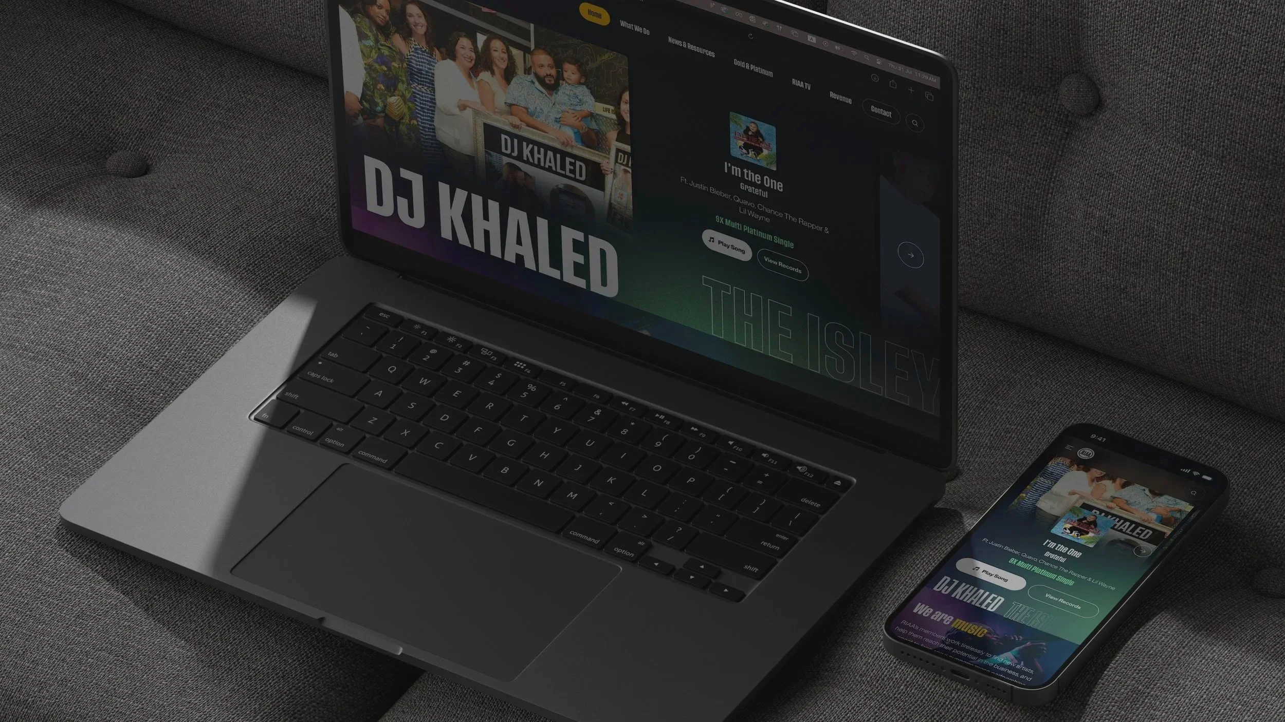 Laptop and smartphone displaying the music artist DJ Khaled's webpage and song, resting on a grey fabric surface.