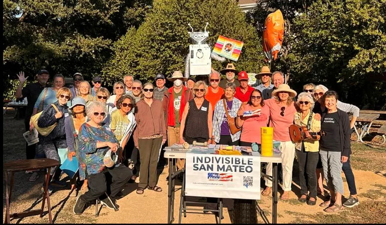 A large group of people gathered outdoors for a group photo, with a table displaying a sign that reads 'INDIVISIBLE SAN MATEO,' and a festive, sunny setting with trees in the background.