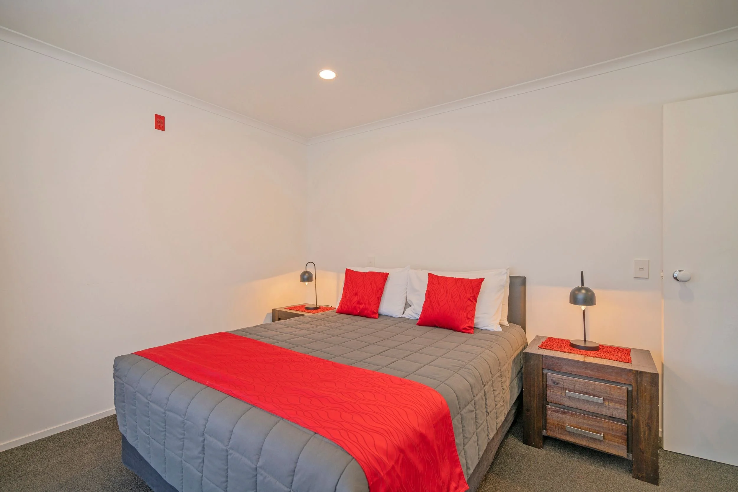 A neatly made bed with gray bedding, red accents, and two nightstands with lamps in a simple, modern bedroom.