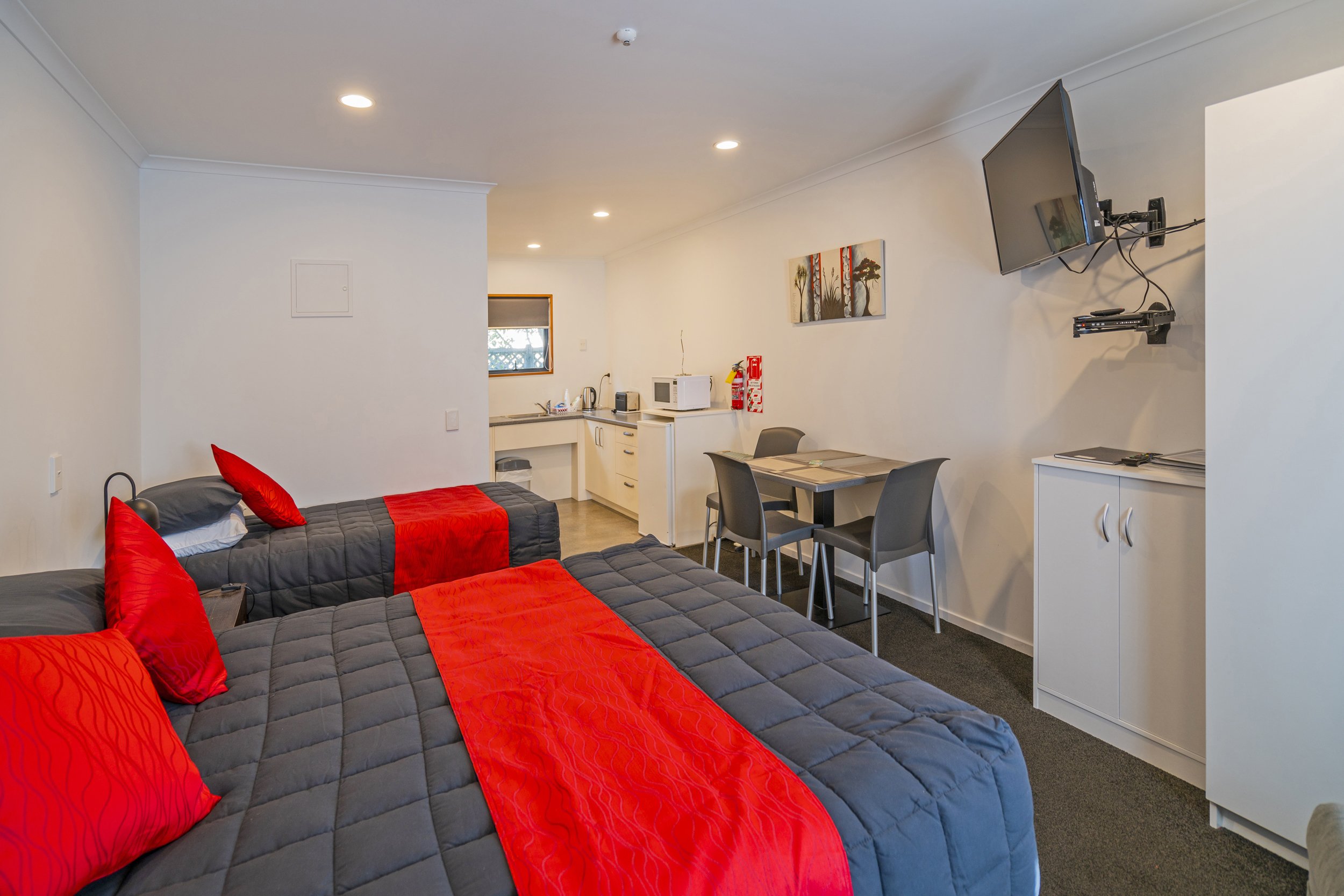 Interior of a small apartment with two beds with red and gray bedding, a kitchenette with small appliances, a dining table with four chairs, a wall-mounted TV, and a white cabinet.