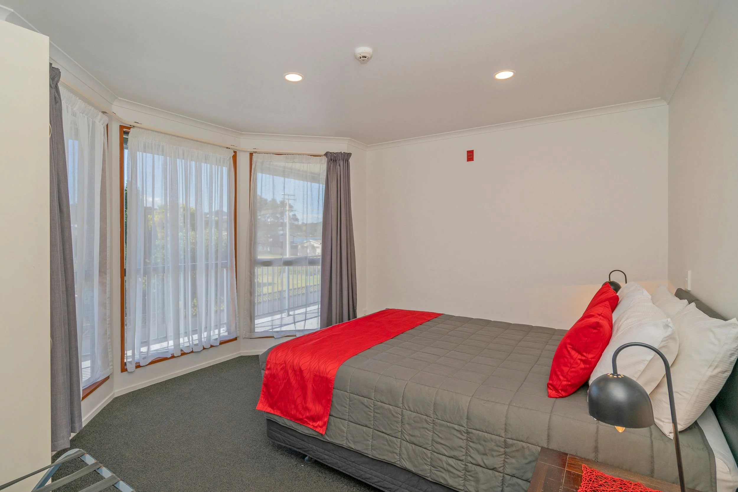 Clean hotel room with large window, white walls, gray bedspread, red accent pillow and throw blanket, black bedside lamps, and gray curtains.