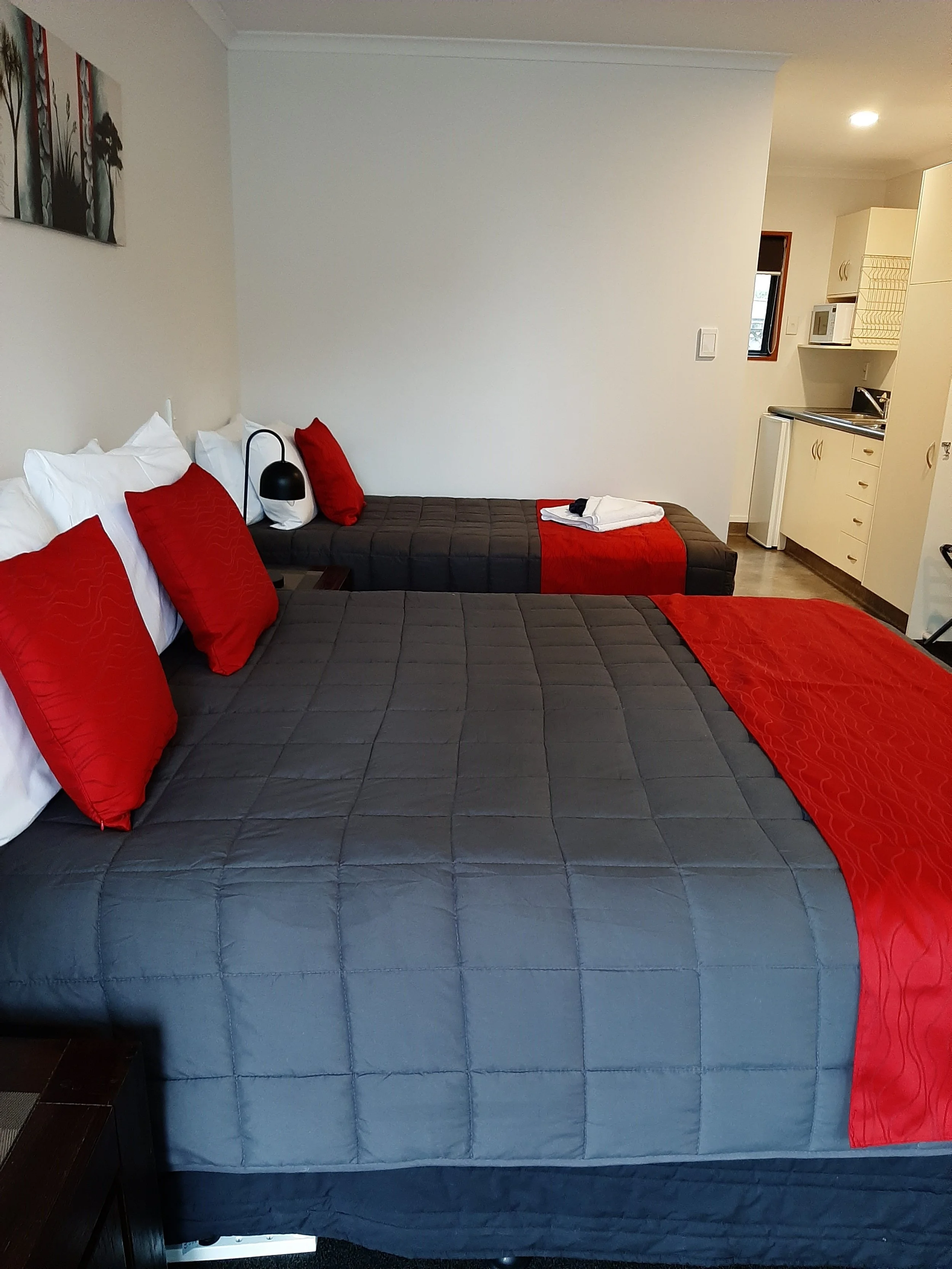 A hotel room with two beds, each with red pillows and a gray bedspread, and a small kitchen area in the background.