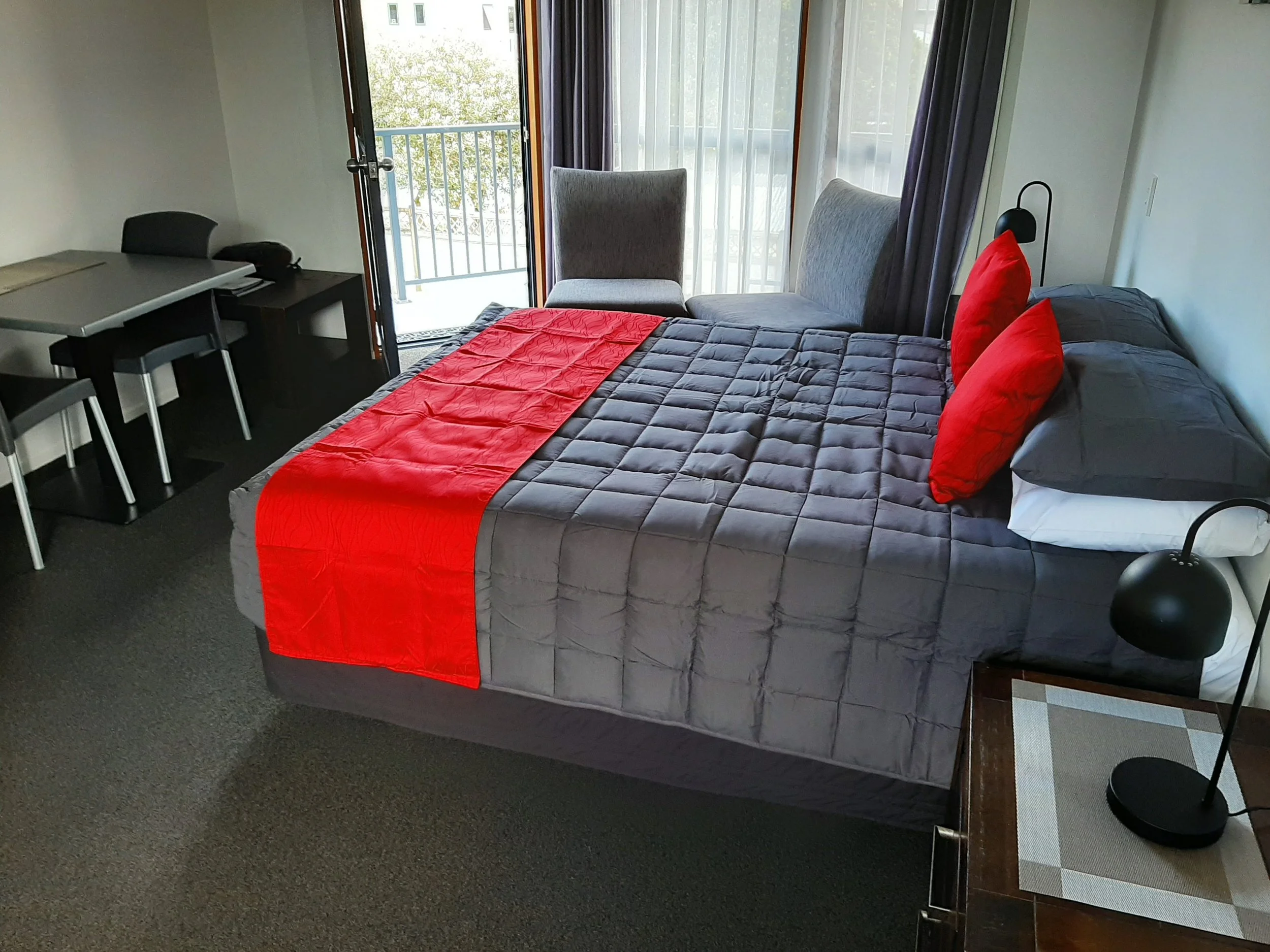 Hotel room with bed featuring gray bedding and red pillows, small desk with chairs, and sitting area near sliding glass door with balcony view.