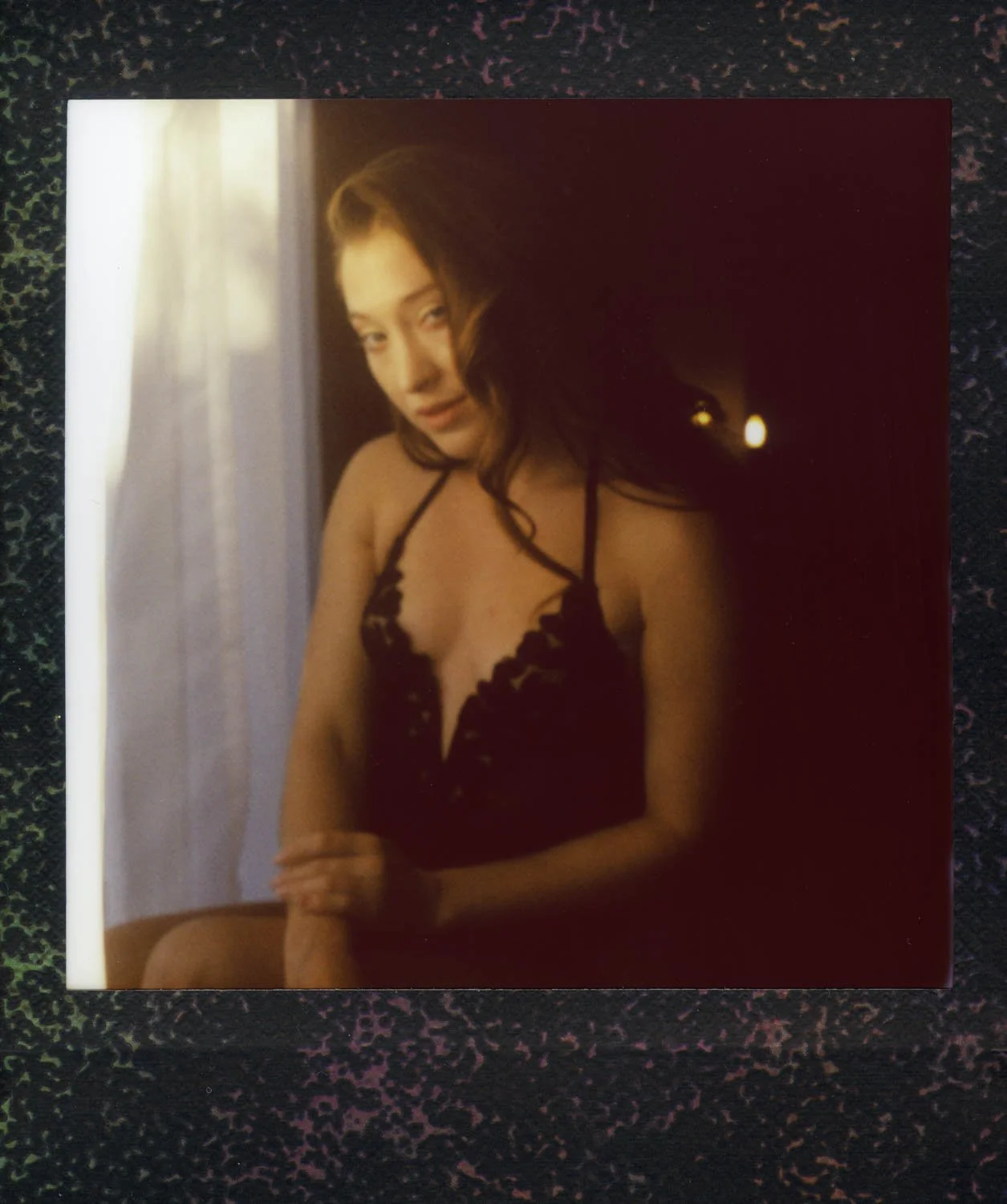 A woman with brown hair in black lace lingerie sitting near a window with sunlight shining through.