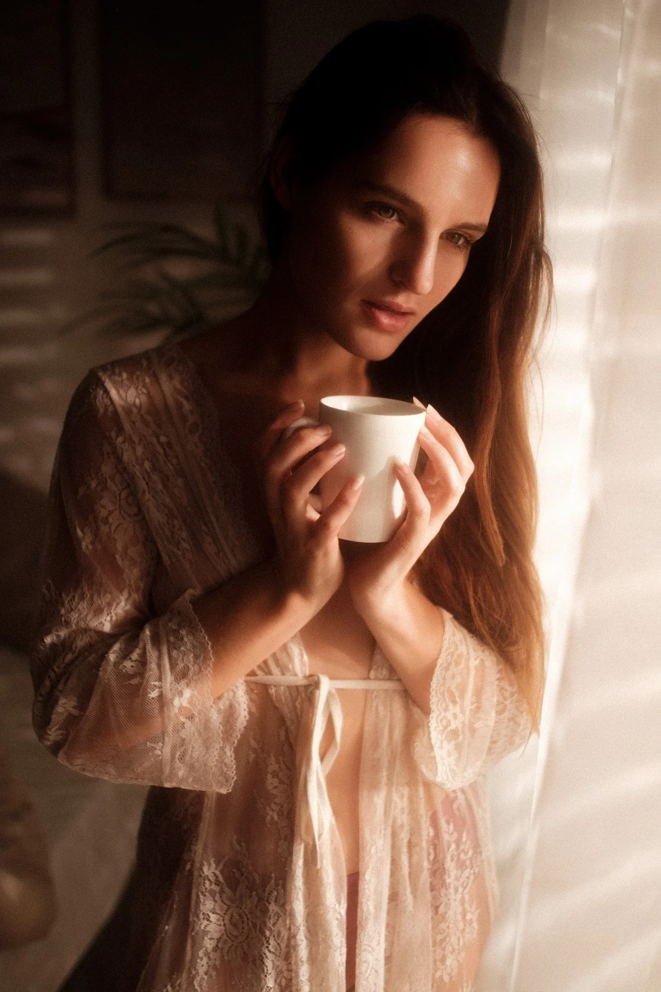 A woman with long brown hair holding a white mug, standing near a light-colored curtain, wearing a lacy, peach-colored robe.