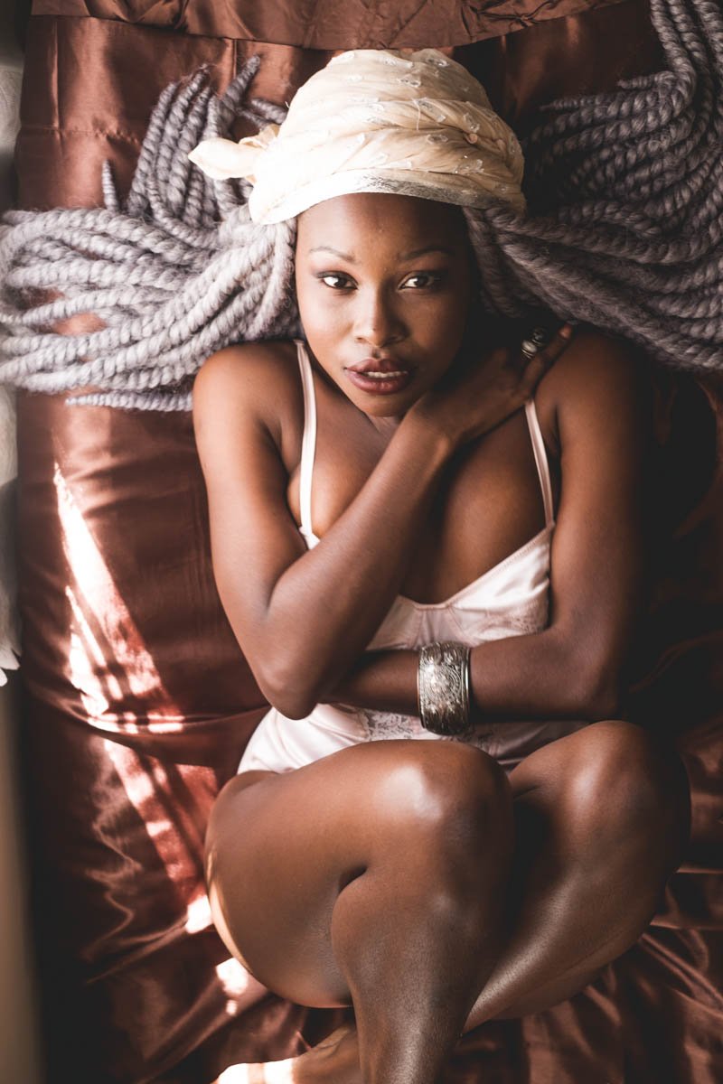 A woman with large gray twisted braids lying on a satin pillow, wearing a white satin slip and a light-colored headscarf, looking directly at the camera.