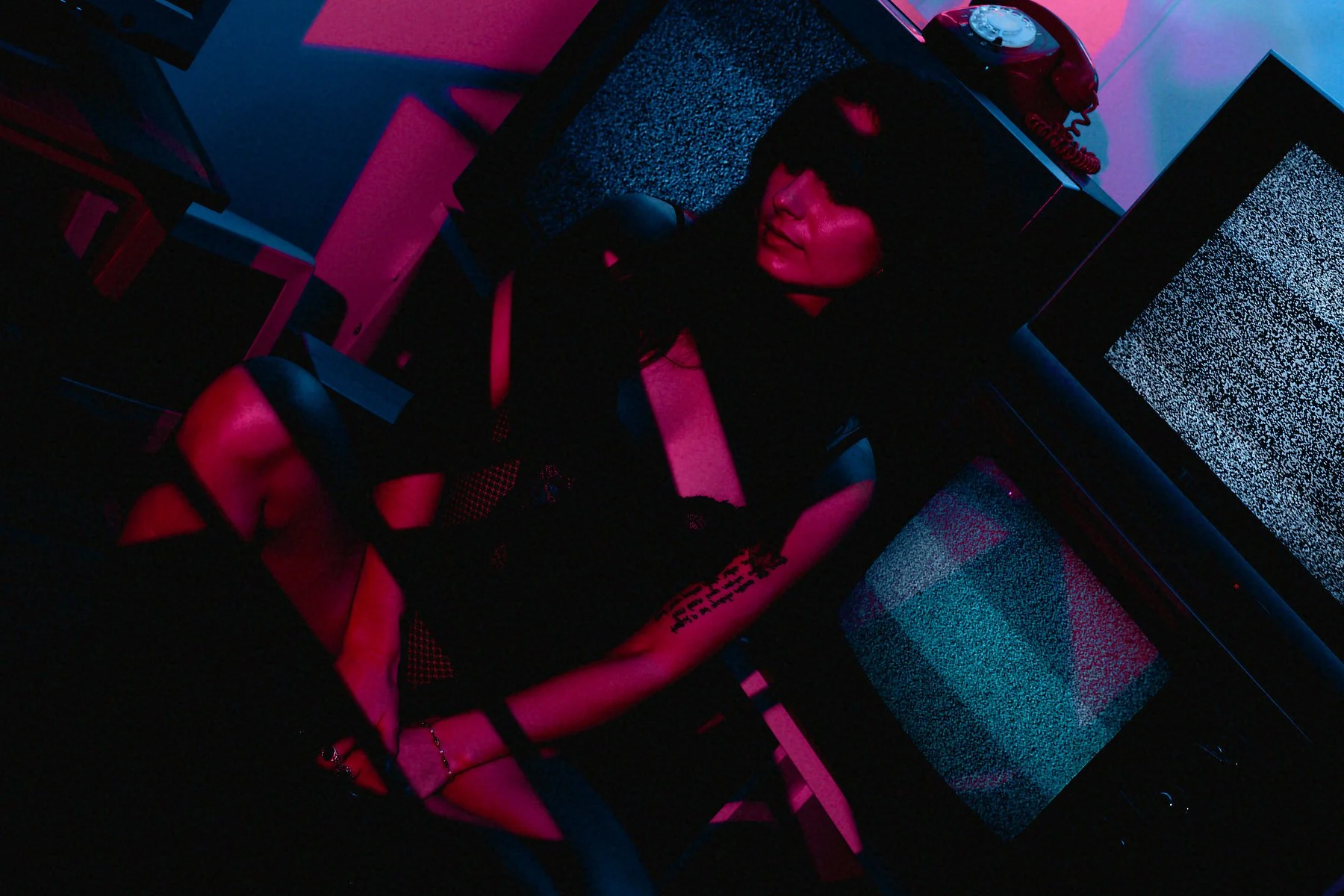 A woman sits at a desk with vintage radio and monitor screens in a dark room illuminated by pink and blue neon lighting, creating a cyberpunk aesthetic.