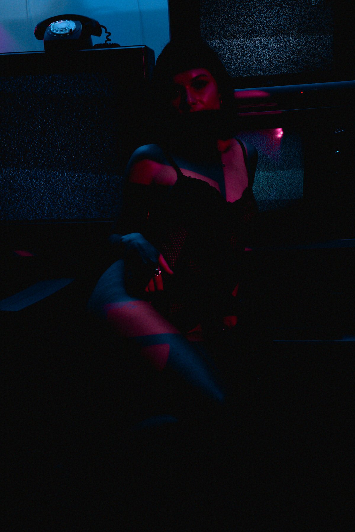 A woman sitting in a dark room illuminated by pink and blue lighting, with an old rotary phone on a table or shelf nearby.