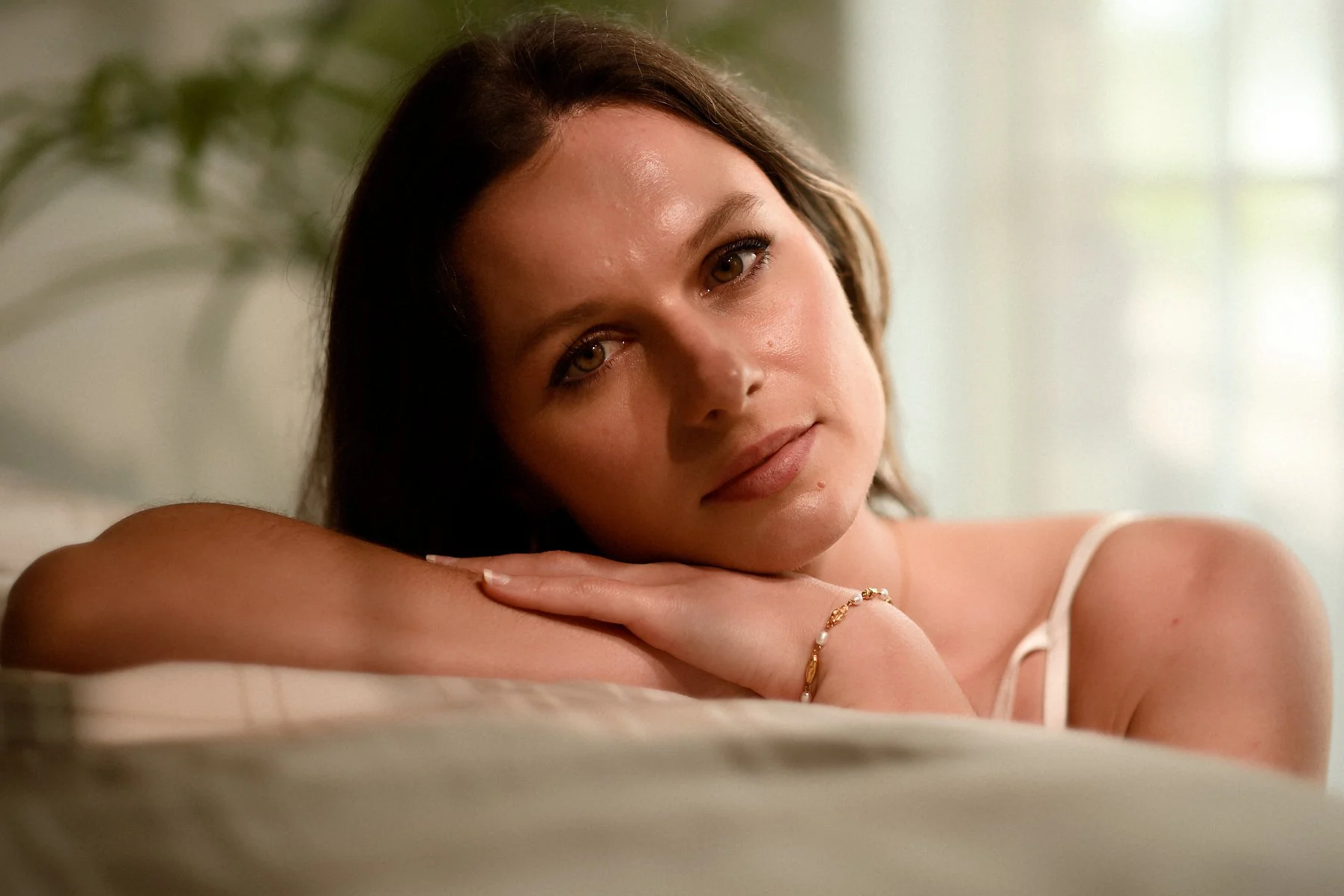 A woman with brown hair resting her head on her arms and looking into the camera, wearing a thin-strapped top and a gold bracelet, with a soft background.