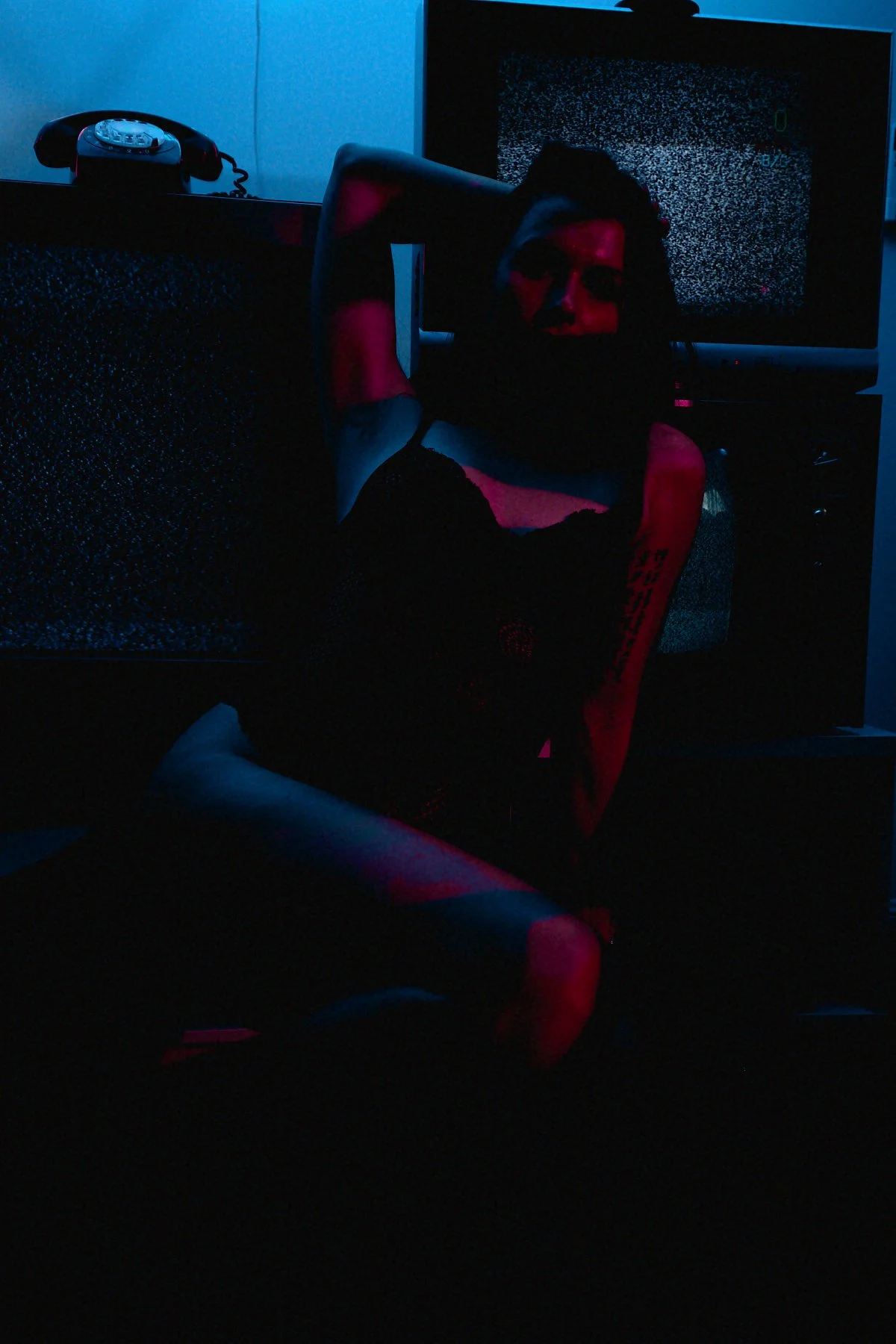 A person sitting in a dark room illuminated by a screen displaying static. The person is partially visible with red lighting on their face and head, and is sitting on a red gaming chair with their arms behind their head. A vintage rotary phone is on 