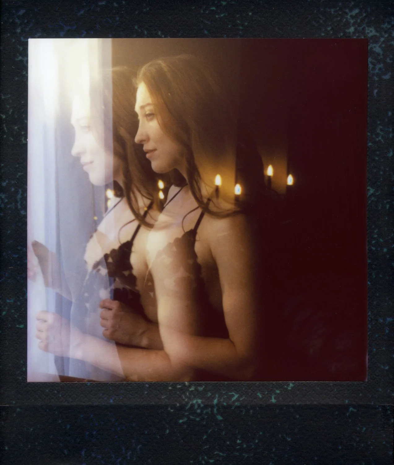 Photo of a woman looking out a window, reflecting another woman holding a flower, in warm light with candles in the background.