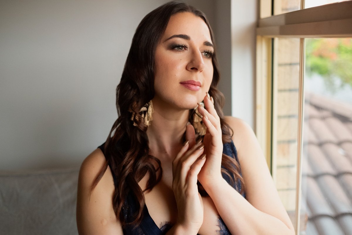 A woman with long brown hair and earrings gazing thoughtfully out a window with a serene expression.