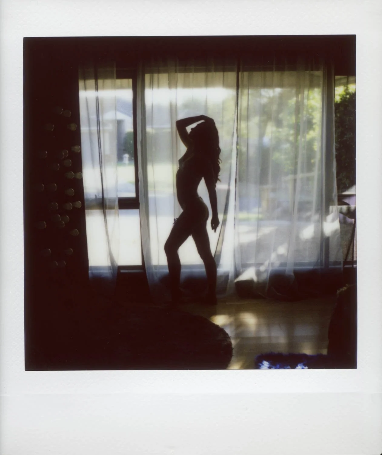 Silhouette of a woman standing in front of a large window with curtains, outside view of trees and a house, sunlight illuminating the scene.