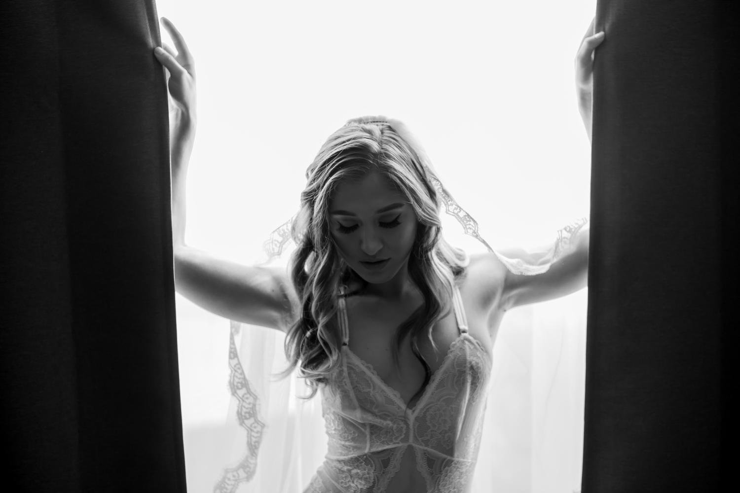 A woman with wavy hair wearing bridal lingerie, standing between curtains, looking downward.