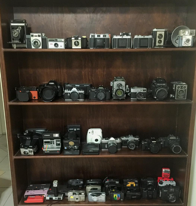 A wooden bookshelf displaying various vintage and modern cameras across four shelves.