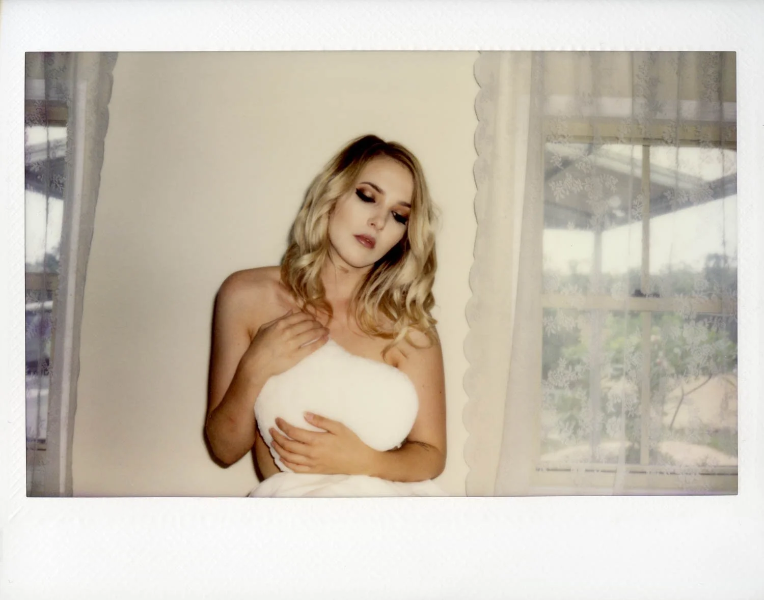 A woman with blonde, wavy hair and makeup, covering her chest with a white towel inside a room with cream walls and sheer curtains, with a view of a balcony and trees outside.