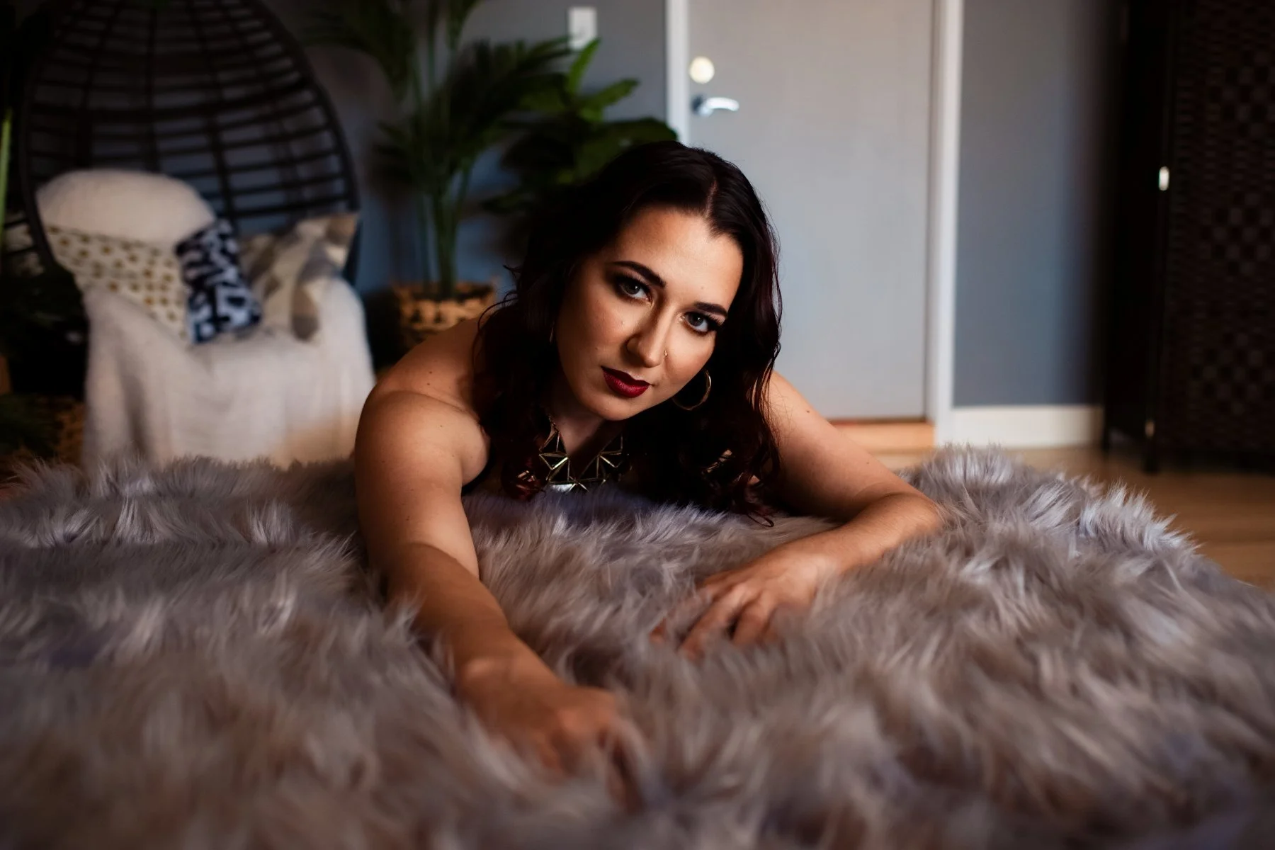 A woman with dark hair and makeup lying on a plush, gray faux fur rug in a room with a bed, plants, and a closed door in the background. She is looking directly at the camera.