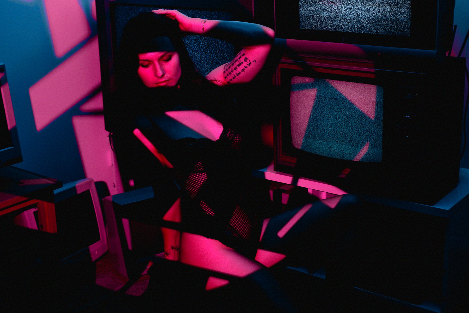 A woman with dark hair and tattoos on her arm is sitting amidst glowing, neon-colored, abstract geometric sculptures, with a television displaying static behind her. The scene is illuminated with vibrant pink and blue lighting.