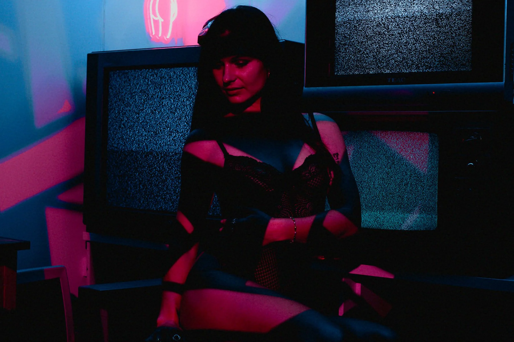 A woman wearing black lingerie, sitting in a dark room illuminated by pink and blue neon lights. She has short dark hair and is surrounded by two TV screens displaying static.